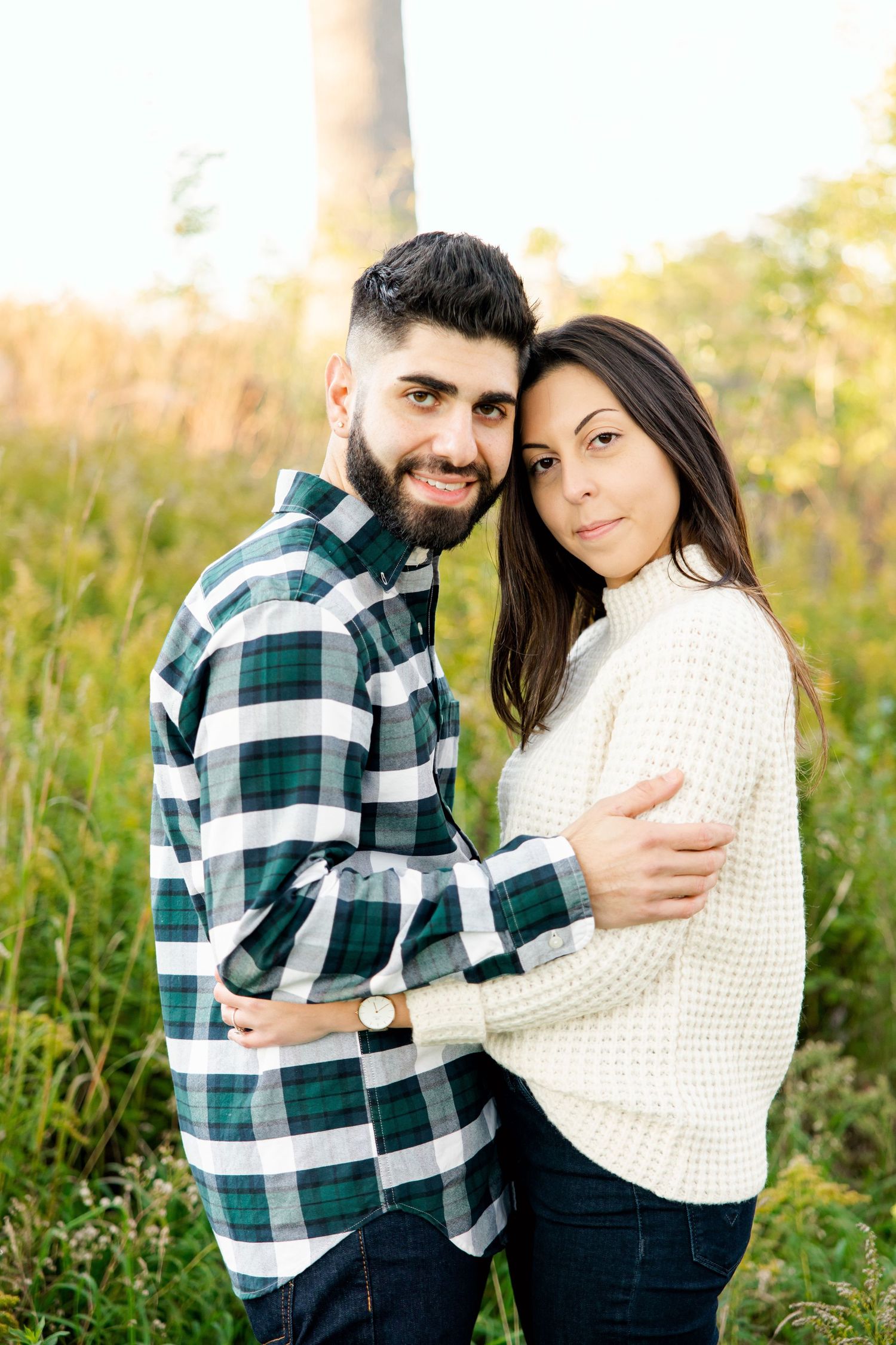 Couple in coordinating plaid shirt and cream sweater embrace while posing in sunny field.