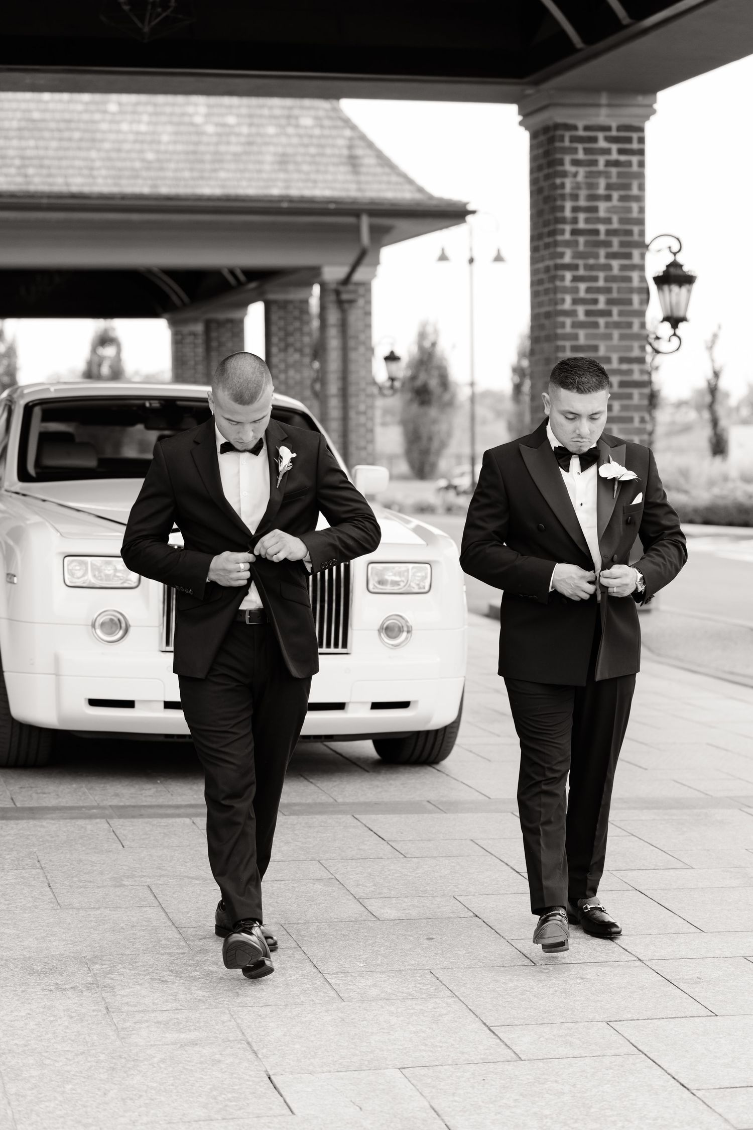 Five wedding party members in black suits walk in a sequence on a stone patio next to a white luxury car.