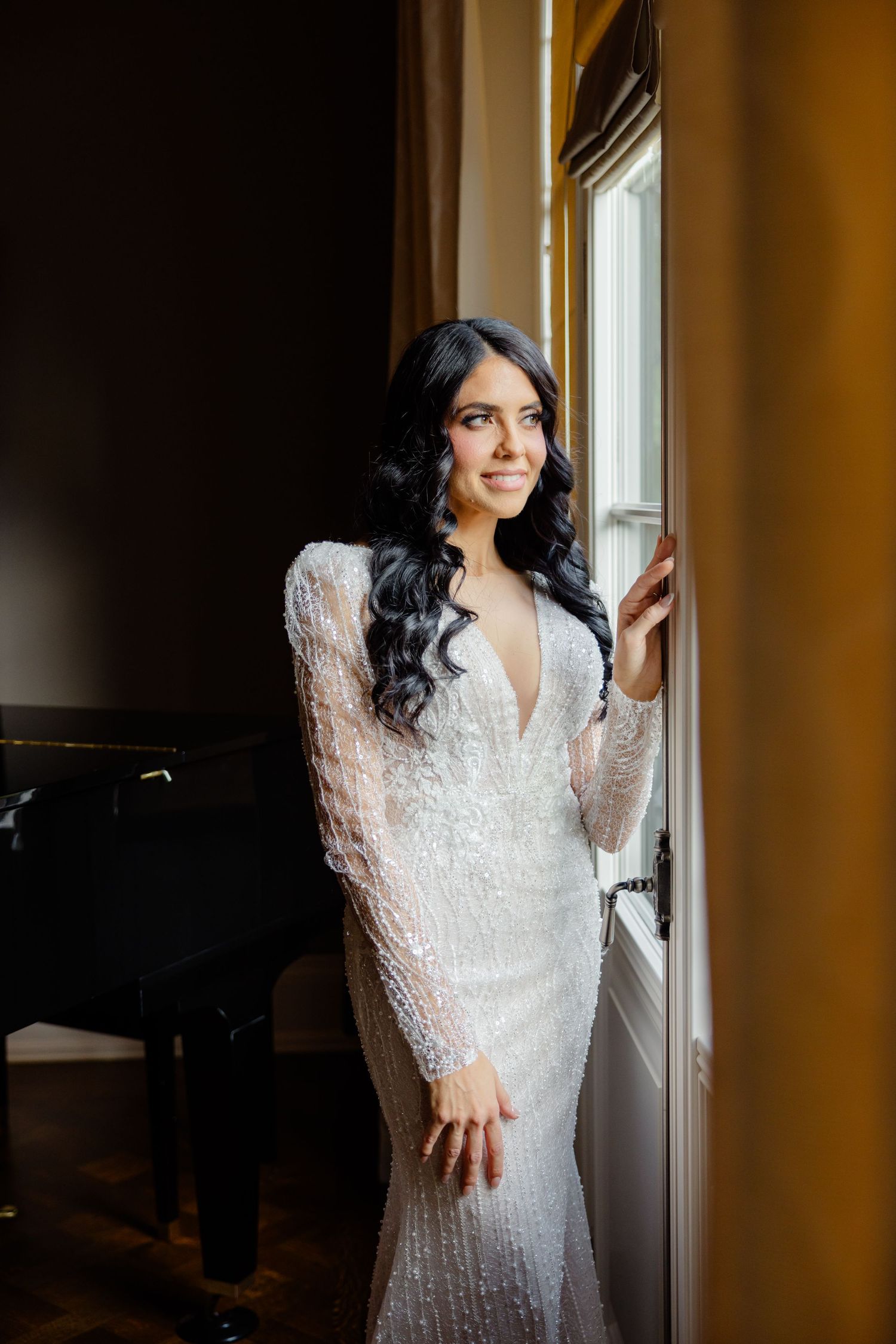 Bride in long sleeve beaded wedding gown poses elegantly by a window in a room with a piano.