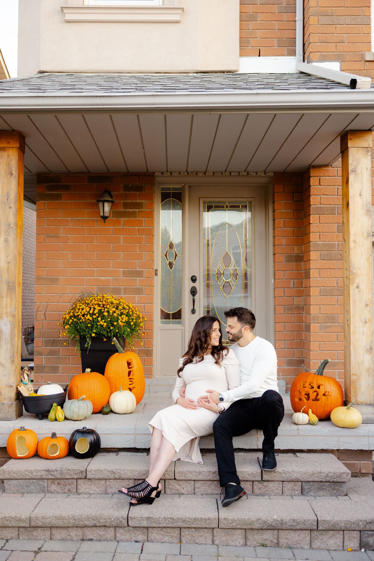 Couple sits on porch steps with autumn decorations including pumpkins and chrysanthemums by a brick house entrance.