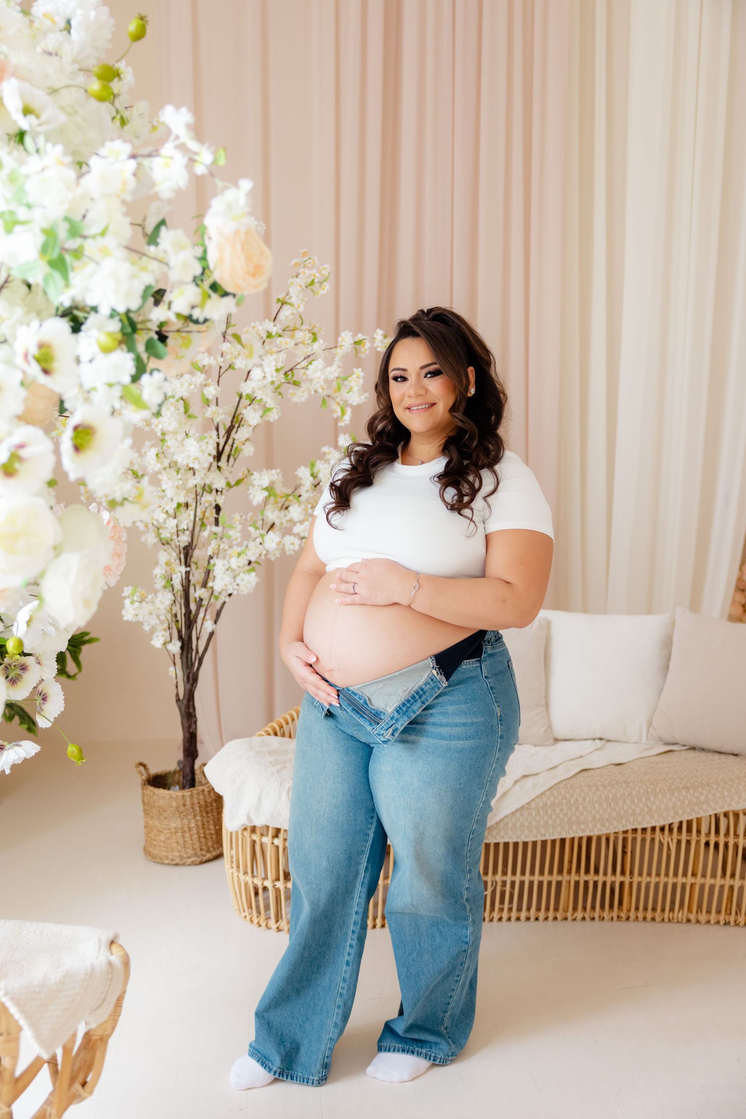 A pregnant woman in a white top and blue jeans poses next to white floral arrangements in a bright studio setting.
