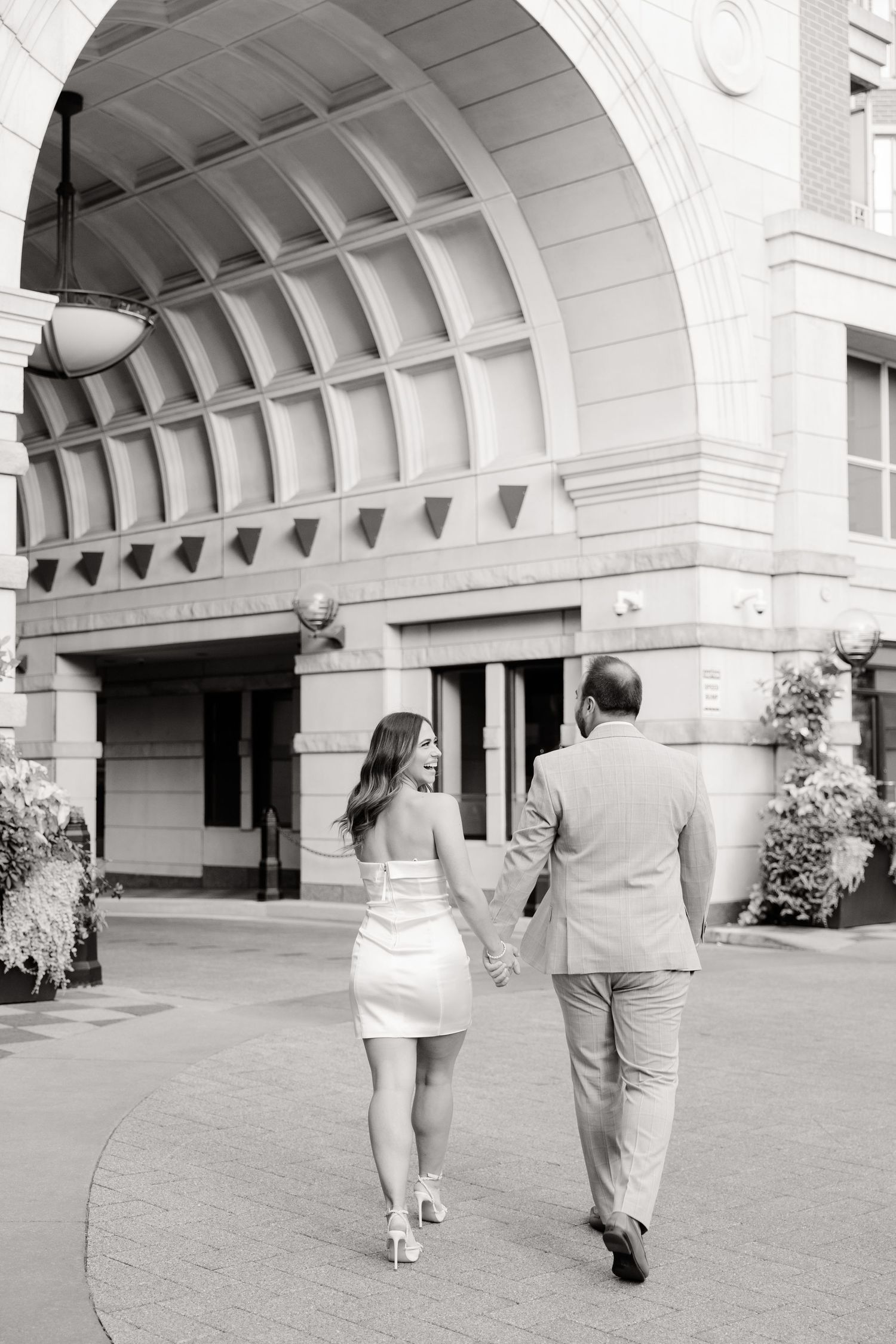 Black and white photo of couple walking under architectural archway holding hands.