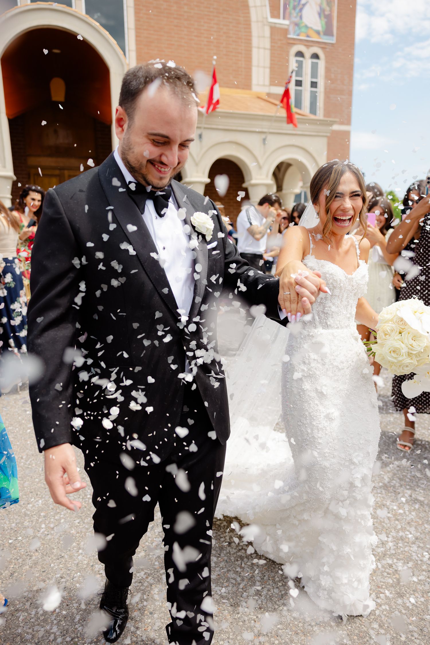 A joyful wedding couple exits their ceremony surrounded by flying white confetti outside a church on a sunny day.