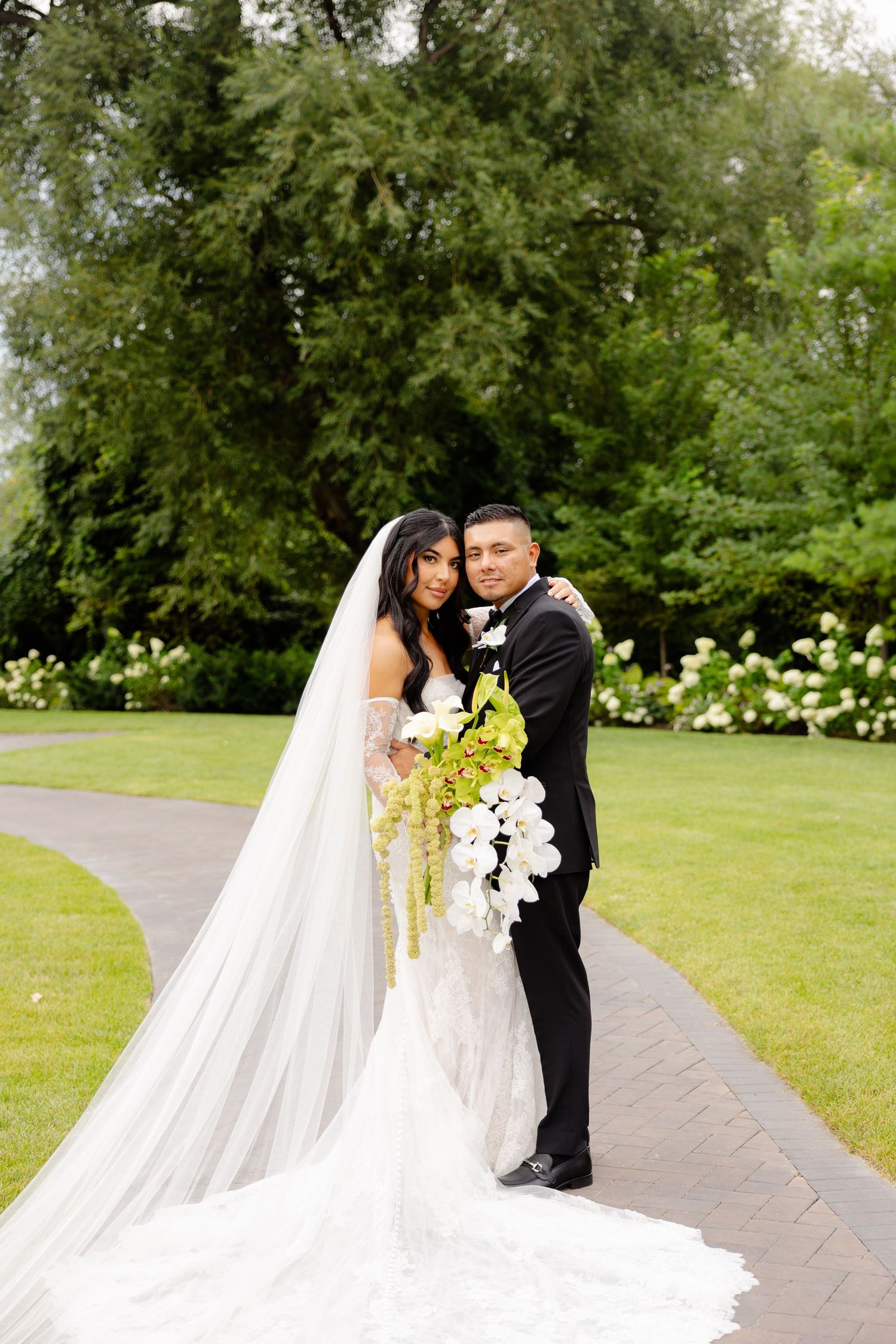 A romantic wedding photo on a garden path with lush greenery and white roses, featuring a flowing white bridal gown.