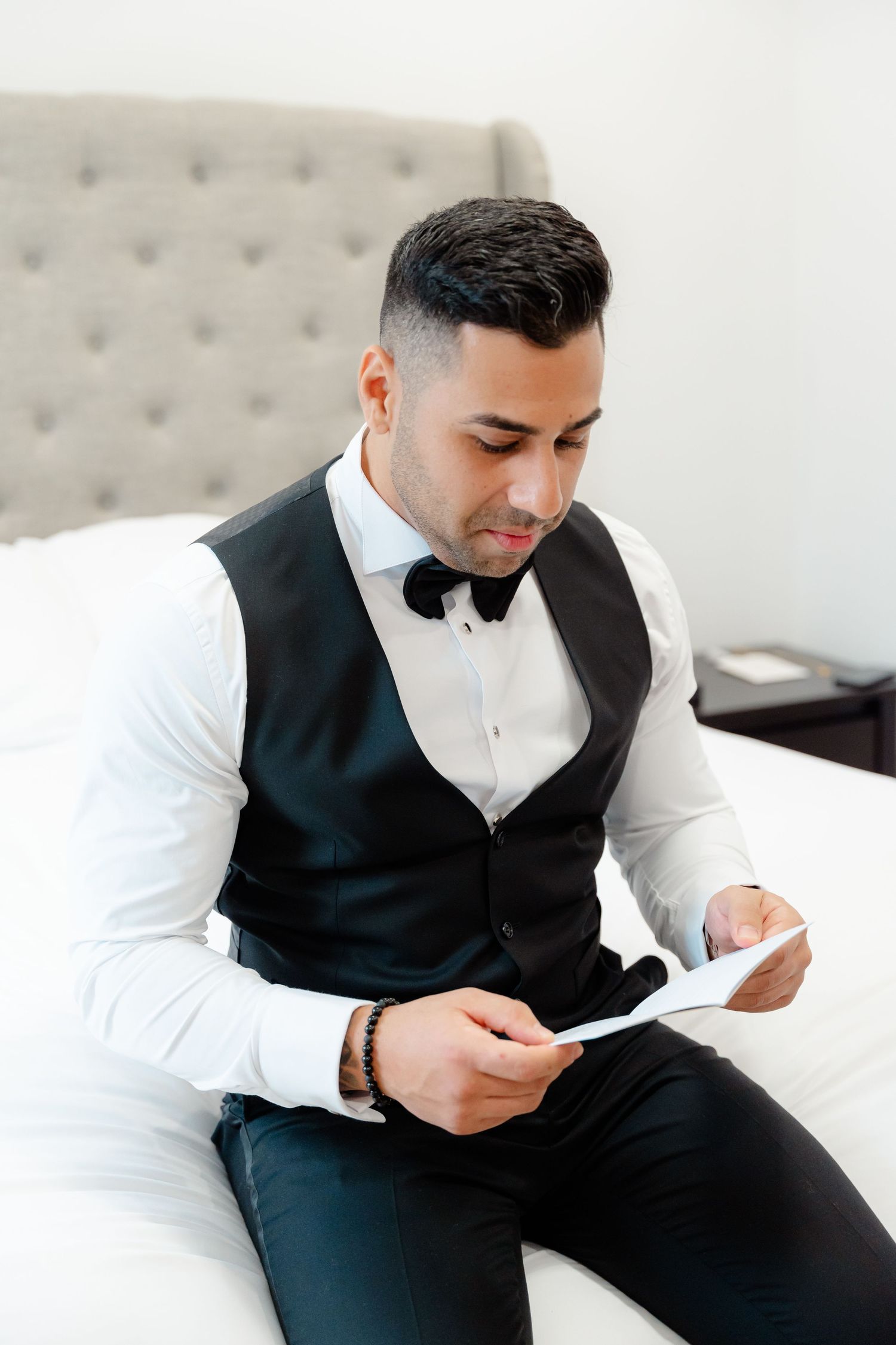 Person in formal vest and bowtie reading a letter while seated on a white couch.