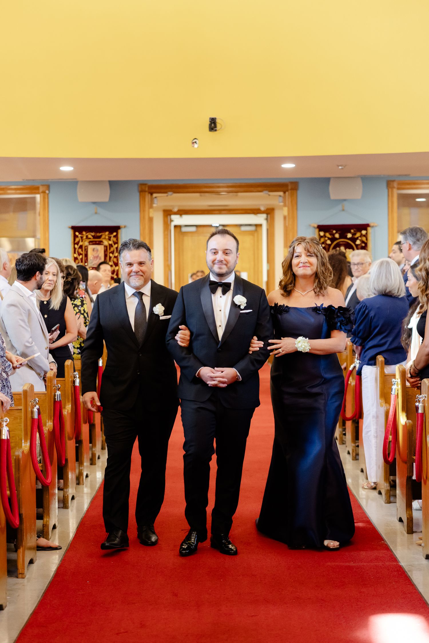 Wedding guests line up along a red carpeted aisle in a church hallway with yellow walls and wooden details.