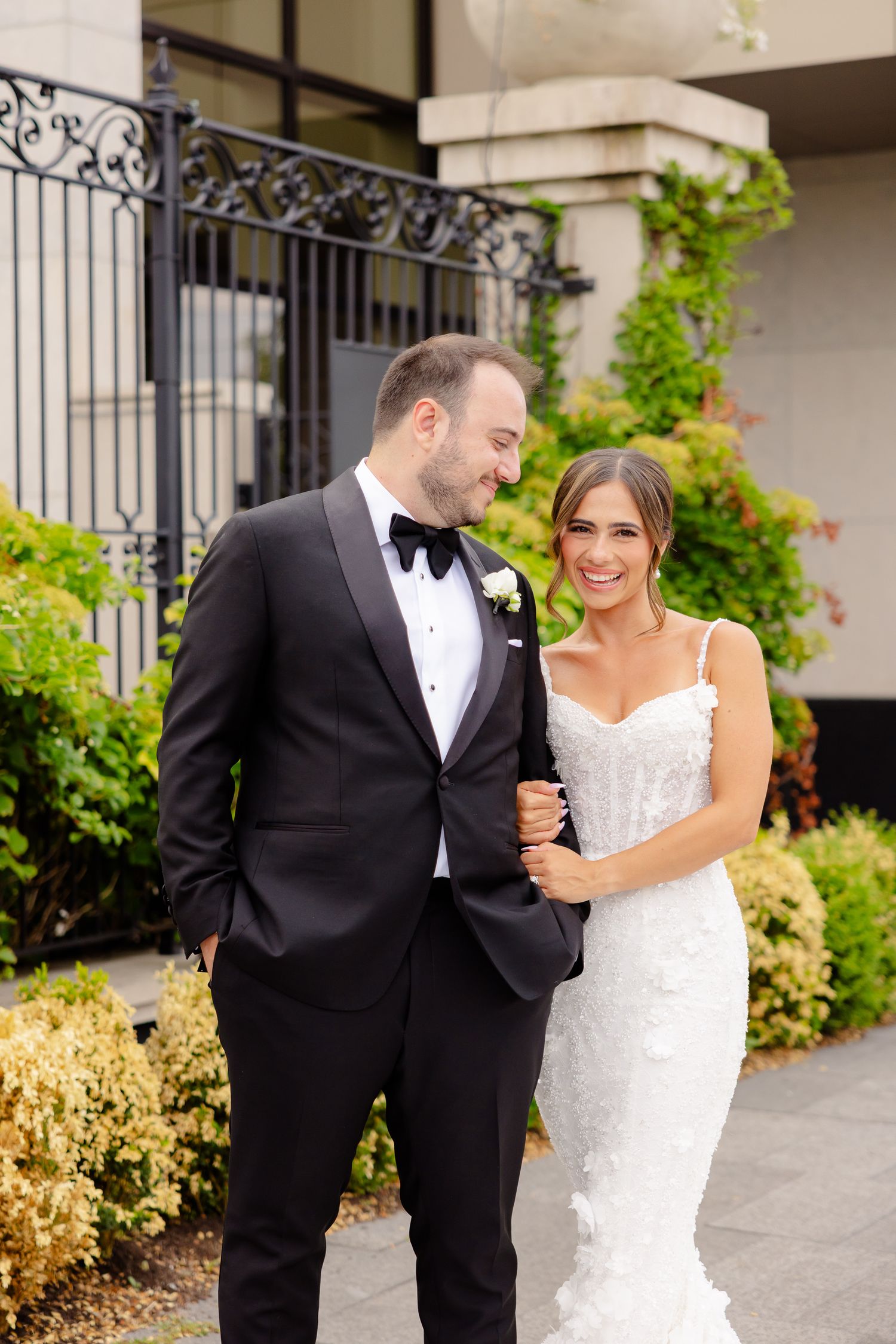 Happy couple share an intimate moment in their wedding attire outside a building with wrought iron gates and greenery.