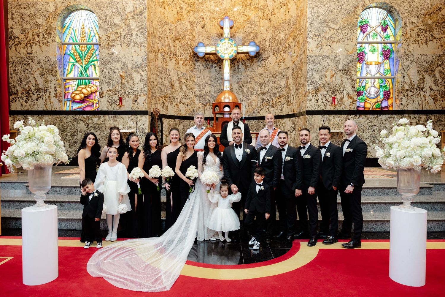 Large wedding party stands in front of church altar with white floral arrangements and stained glass windows.