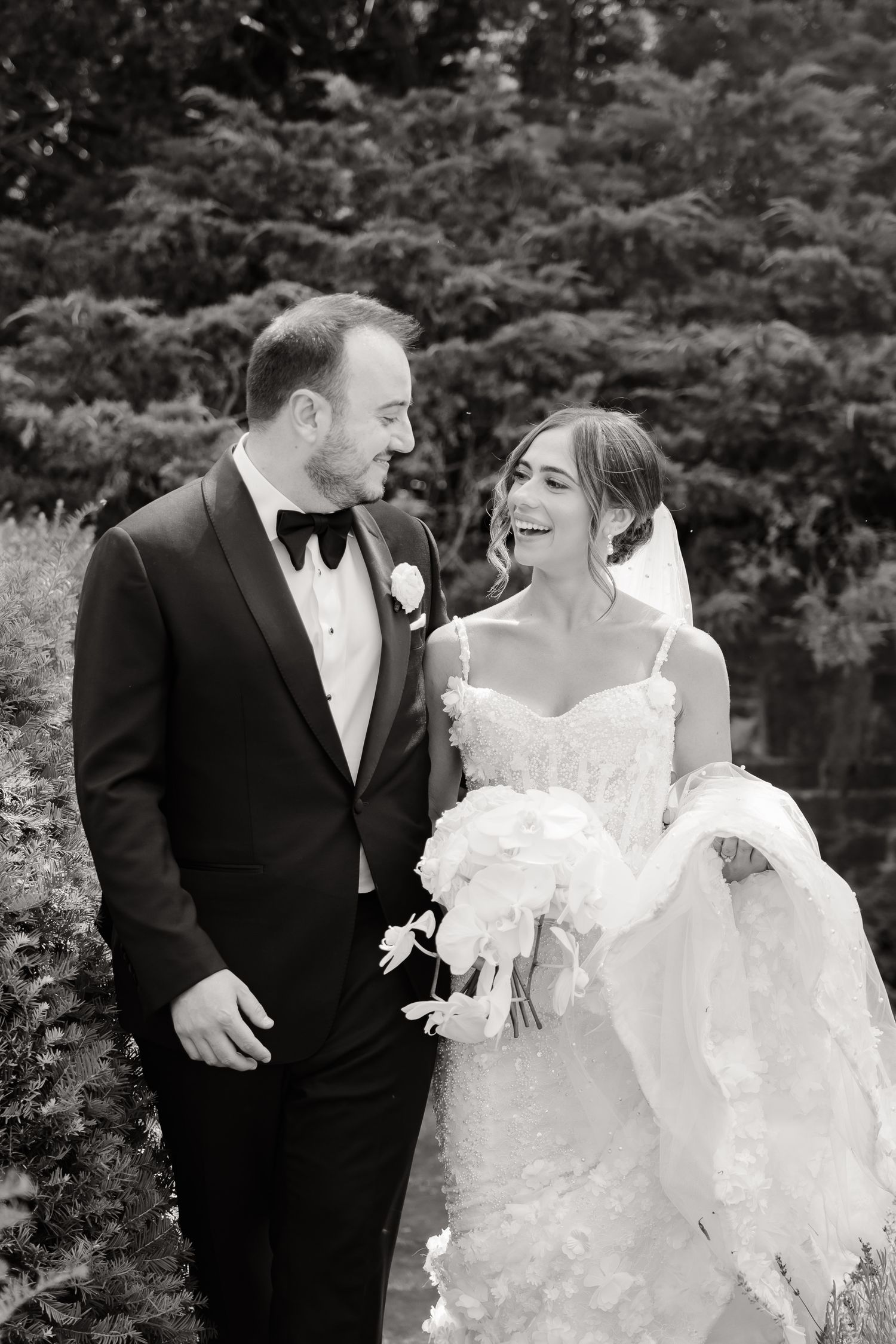 A bride and groom share joyful candid moments together in a garden setting wearing formal wedding attire.