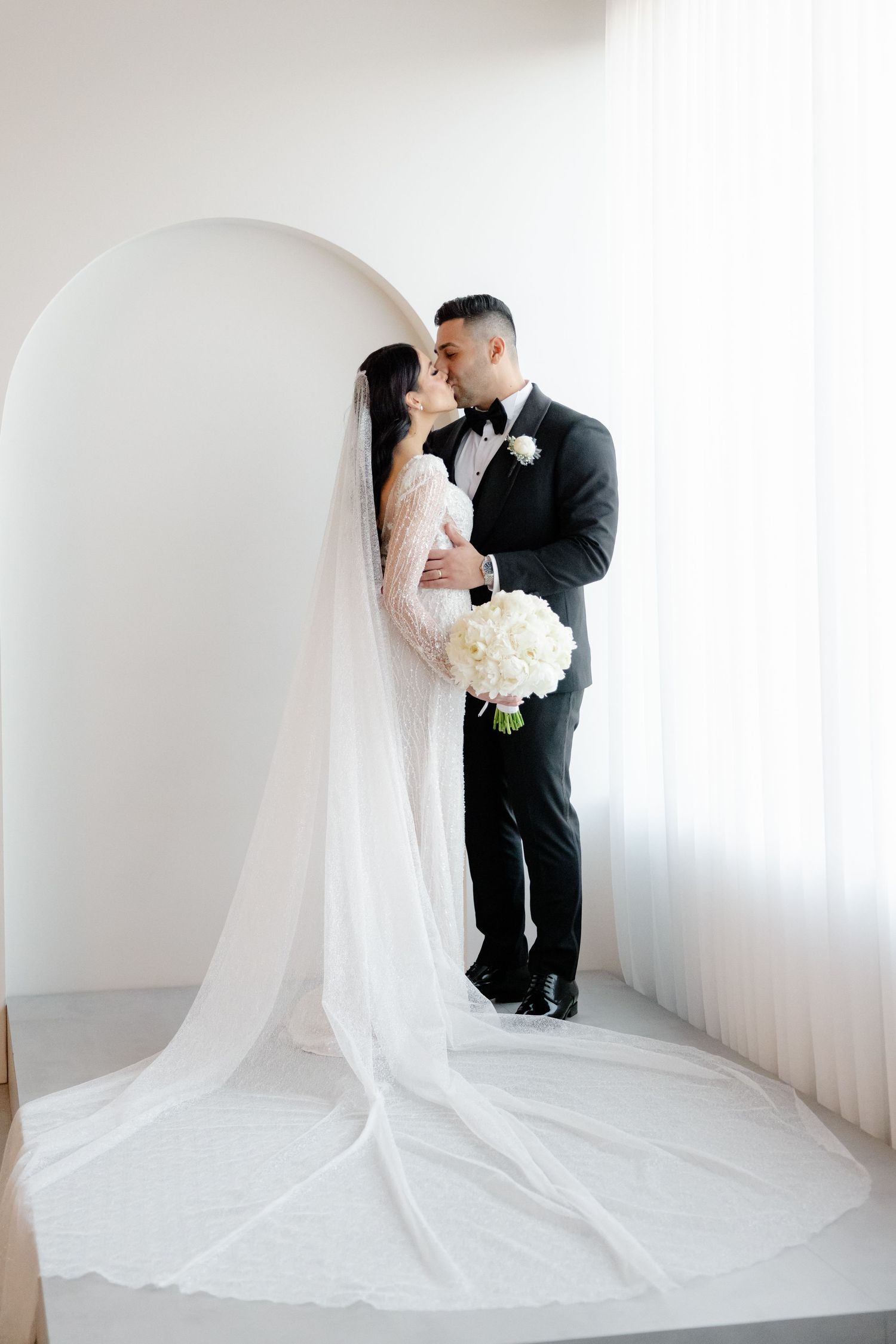 Elegant wedding couple embracing in white archway with dramatic floor-length veil.