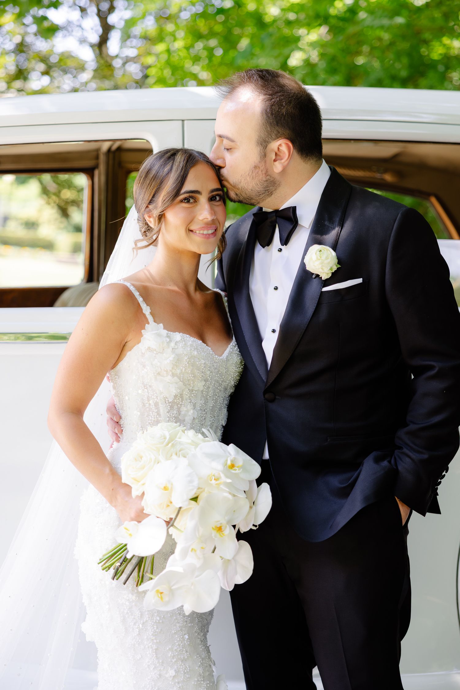 Happy wedding couple stands by vintage white car with beautiful orchid bouquet in elegant formal attire.