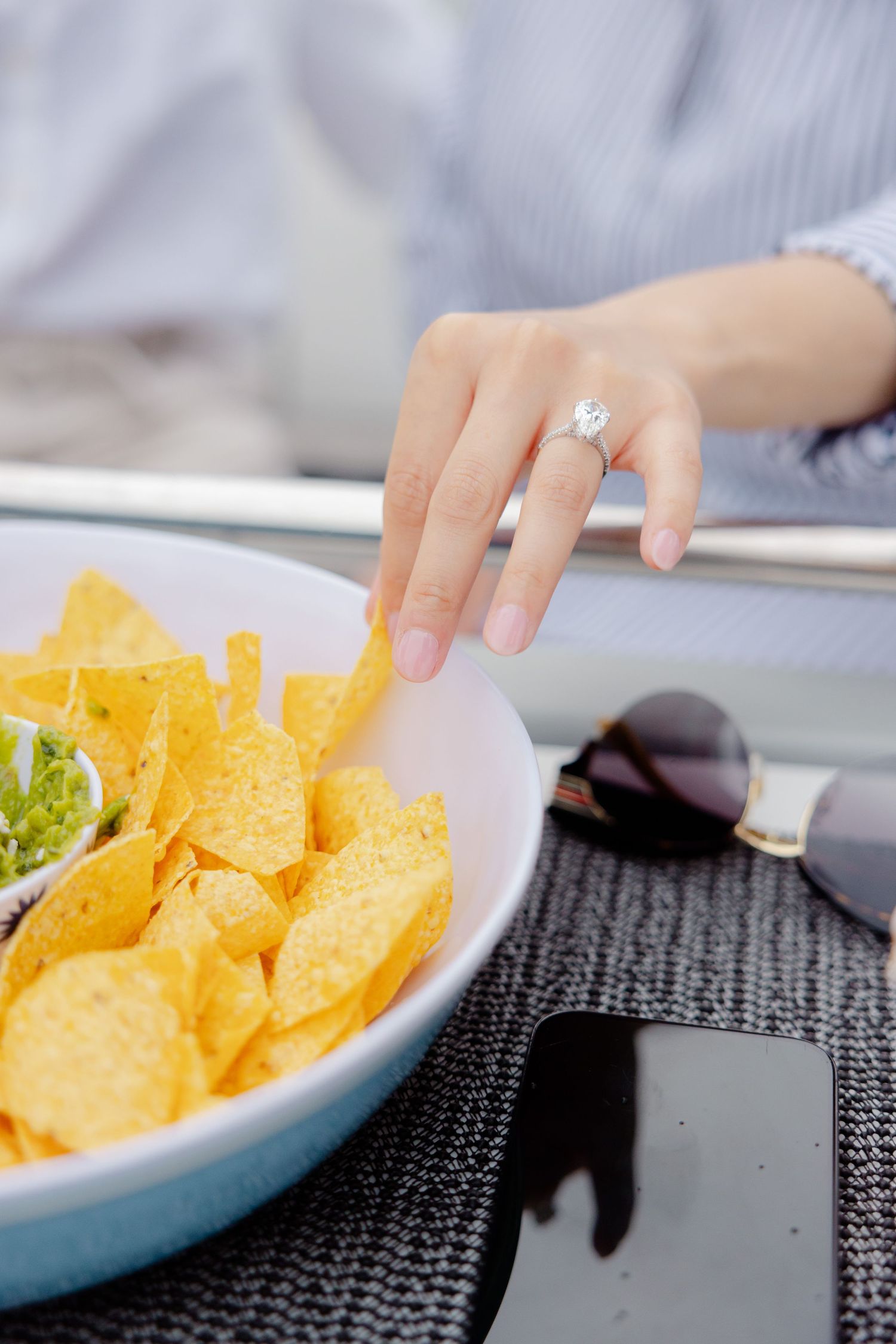 A hand with a diamond ring reaching for yellow potato chips in a blue bowl next to sunglasses and a smartphone.