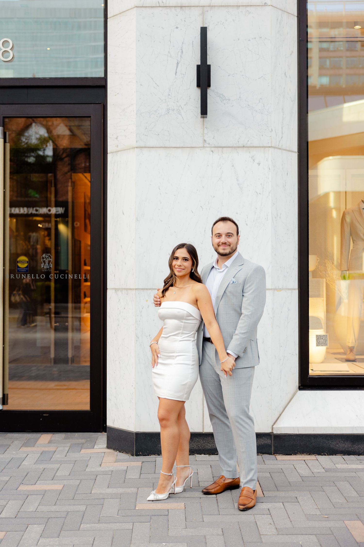 Well-dressed couple poses against modern white marble wall near boutique windows.