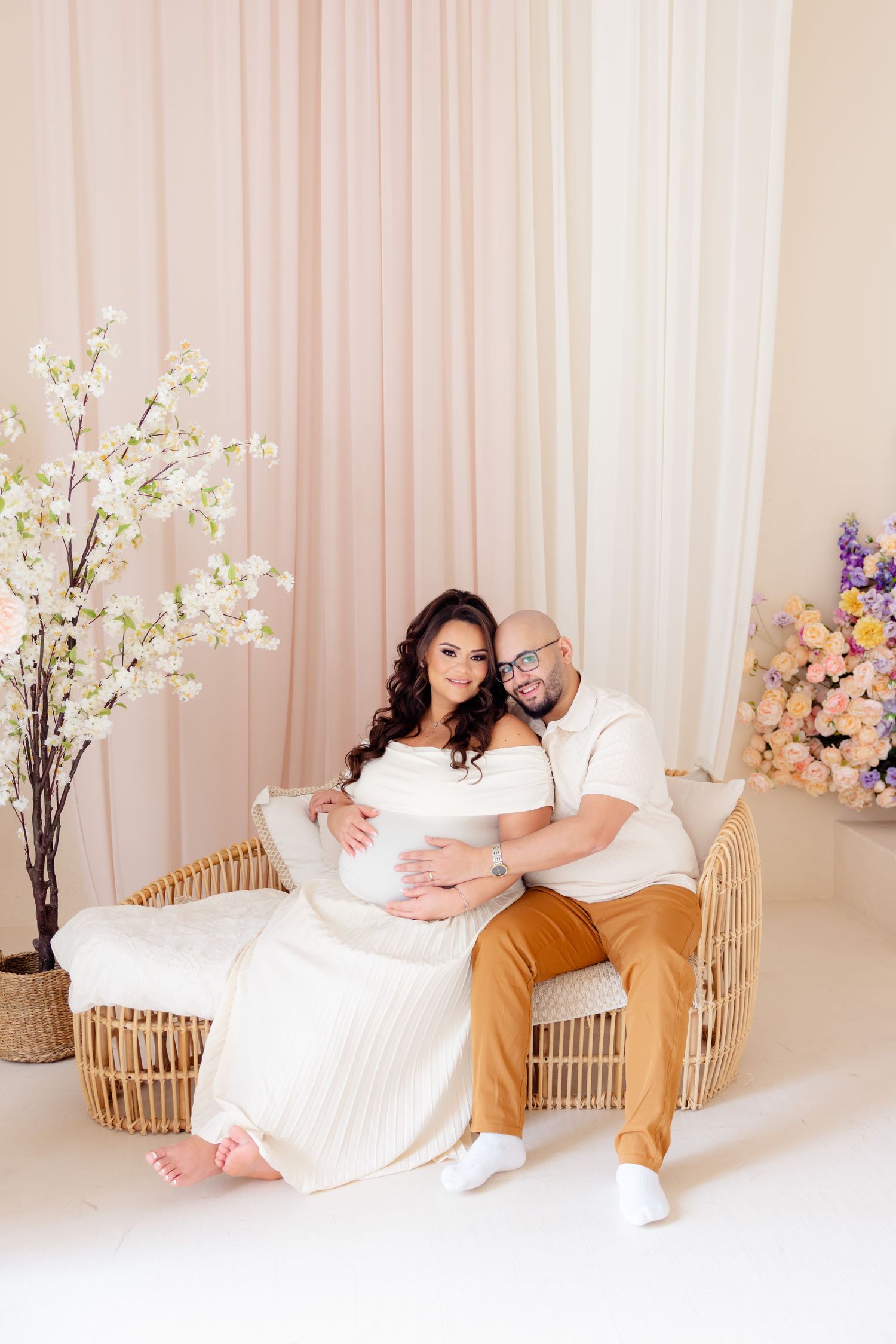 Expectant couple poses together on rattan furniture surrounded by cherry blossoms and floral arrangements in pink studio setting.