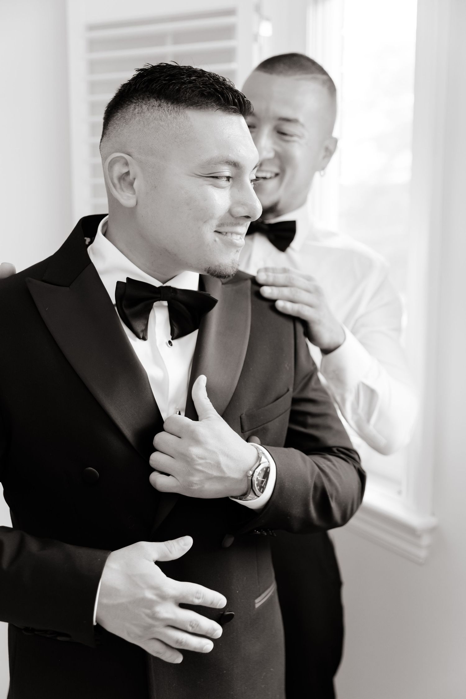 Black and white candid photo of wedding preparations showing a groom in a tuxedo and bow tie smiling with someone behind him.