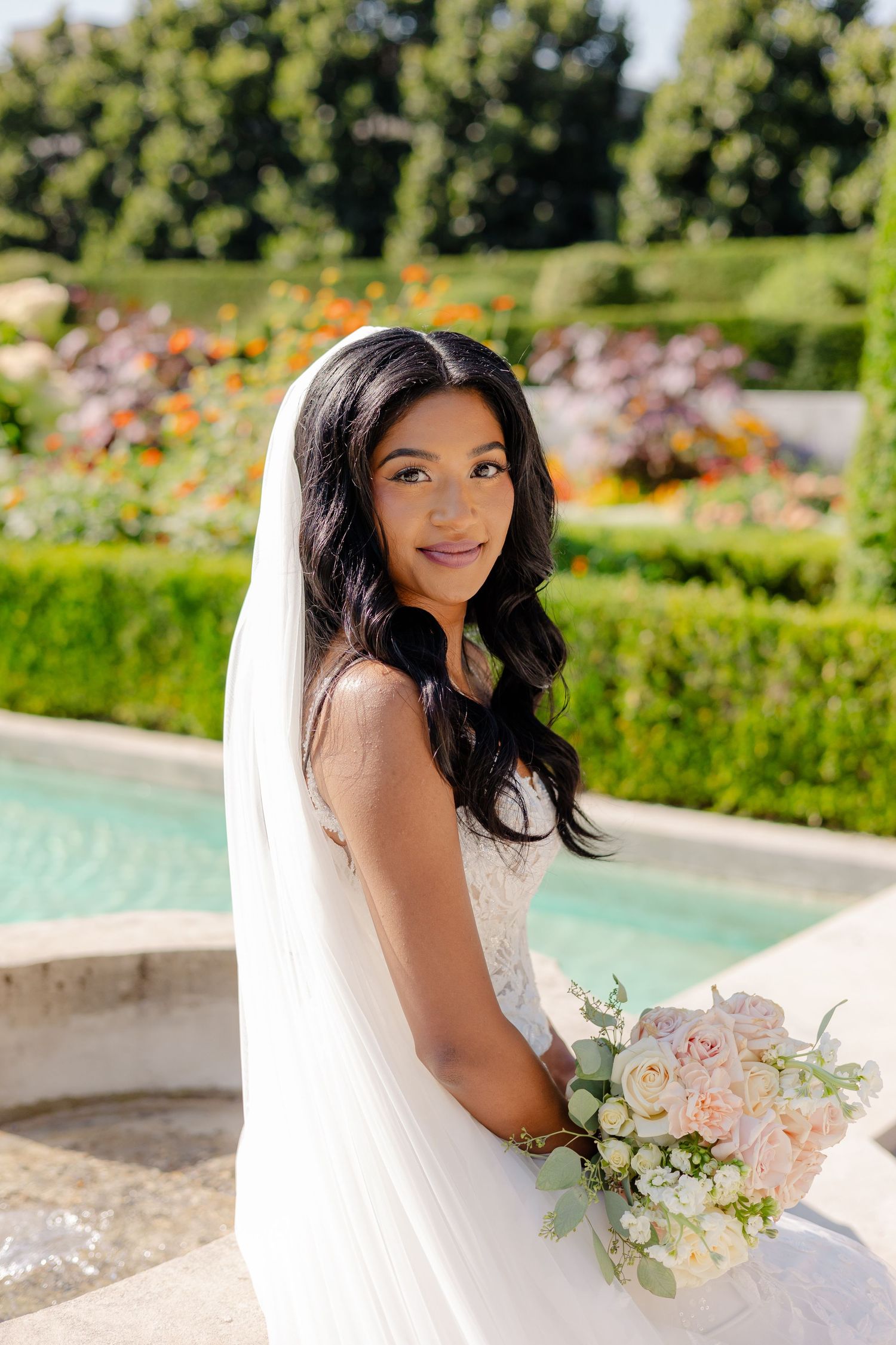 Bride in white lace gown holds pastel bouquet while posing by garden fountain.