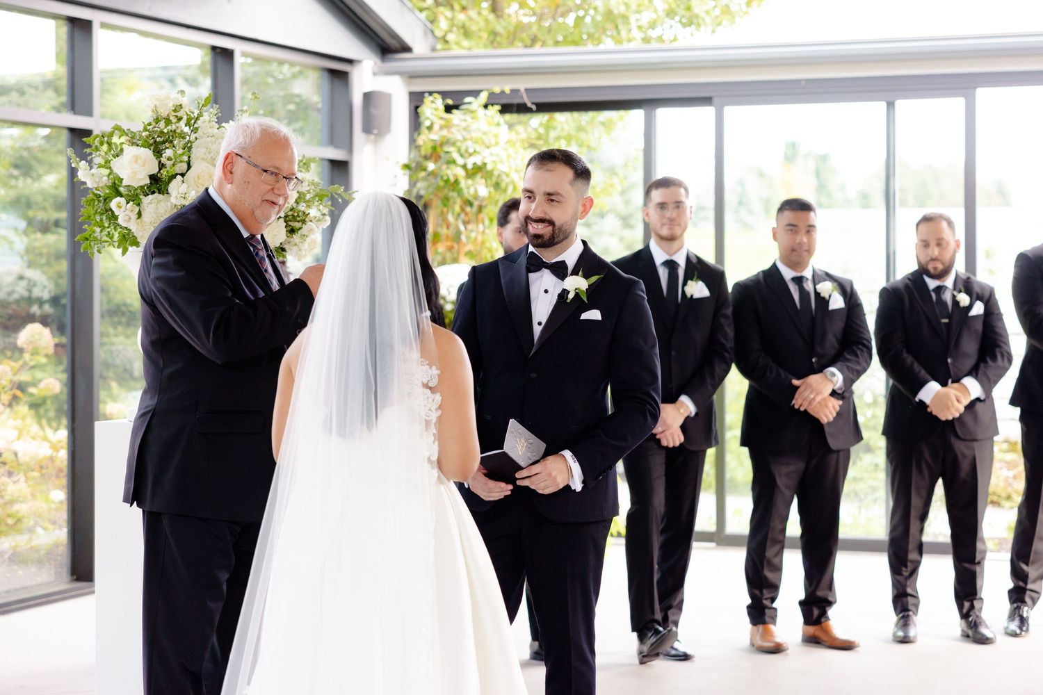 Wedding ceremony in bright venue with groomsmen lined up in black tuxedos.