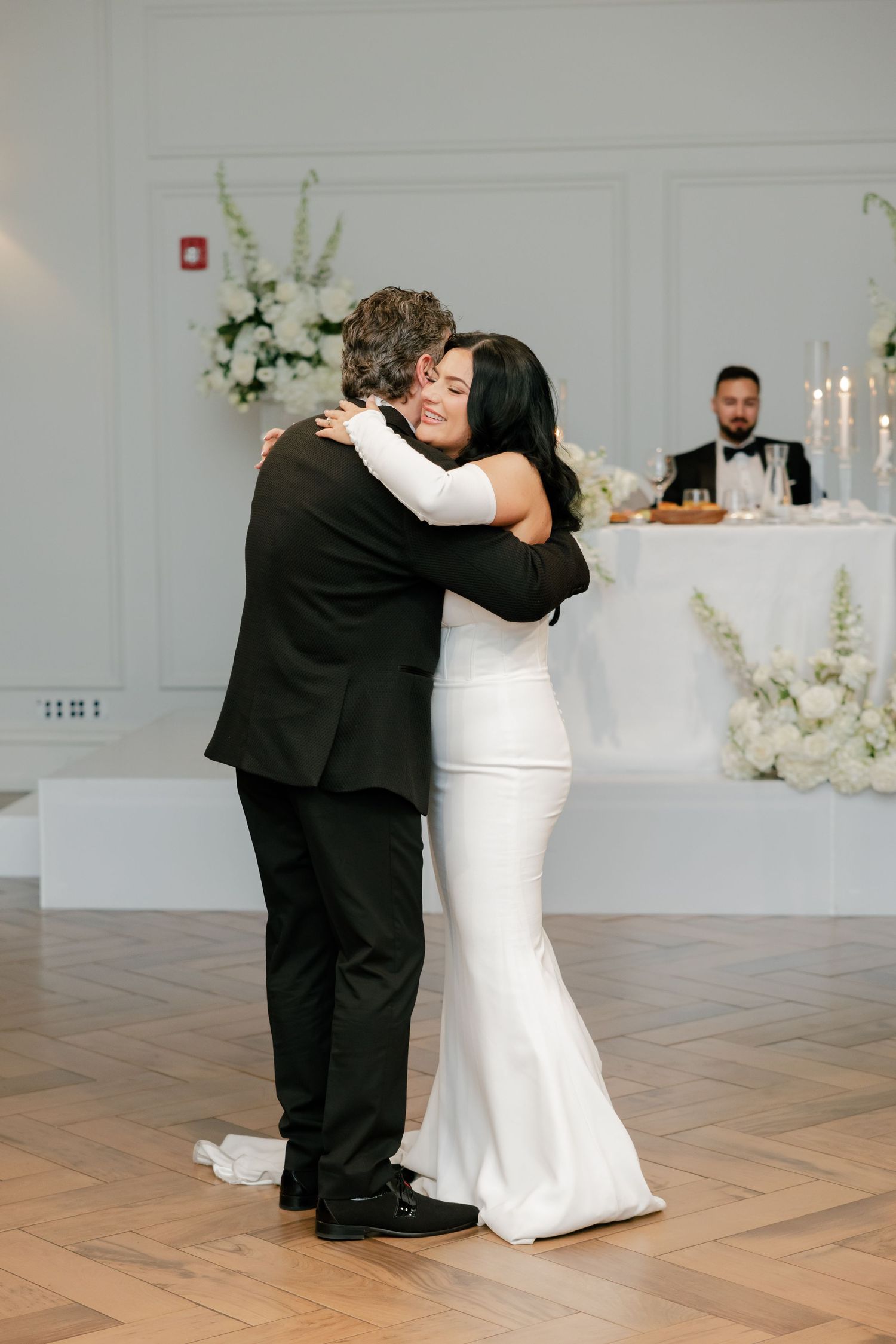 A heartfelt embrace during a wedding dance with elegant white floral decorations in the background.