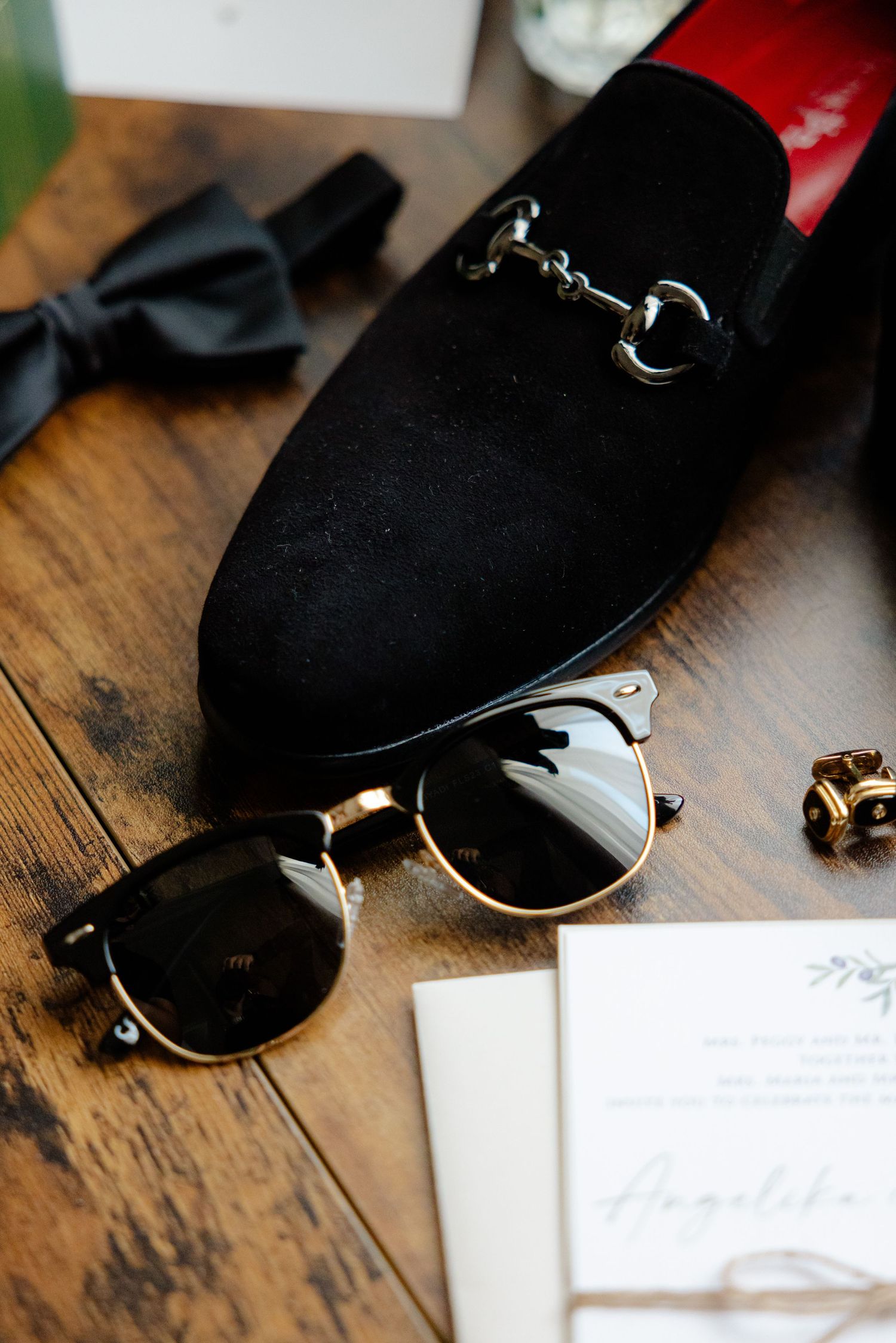 Black suede dress shoes and retro sunglasses next to wedding invitation.