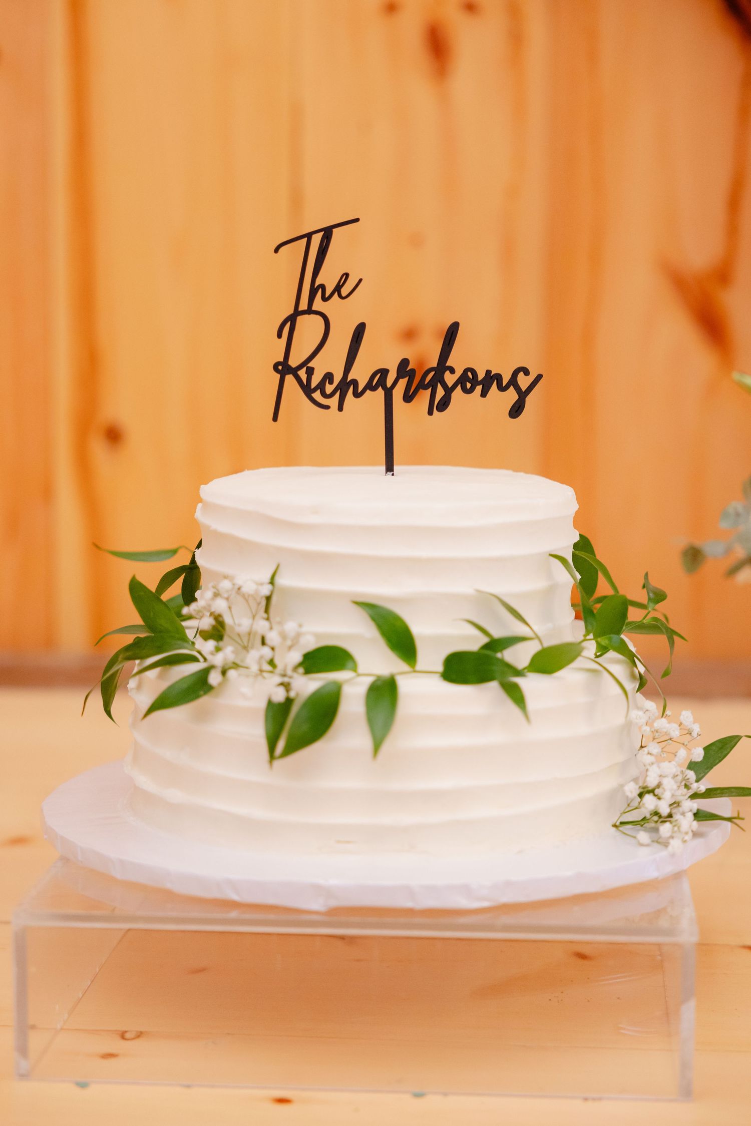 White two-tier wedding cake decorated with greenery and personalized 'The Richardsons' cake topper on clear stand.