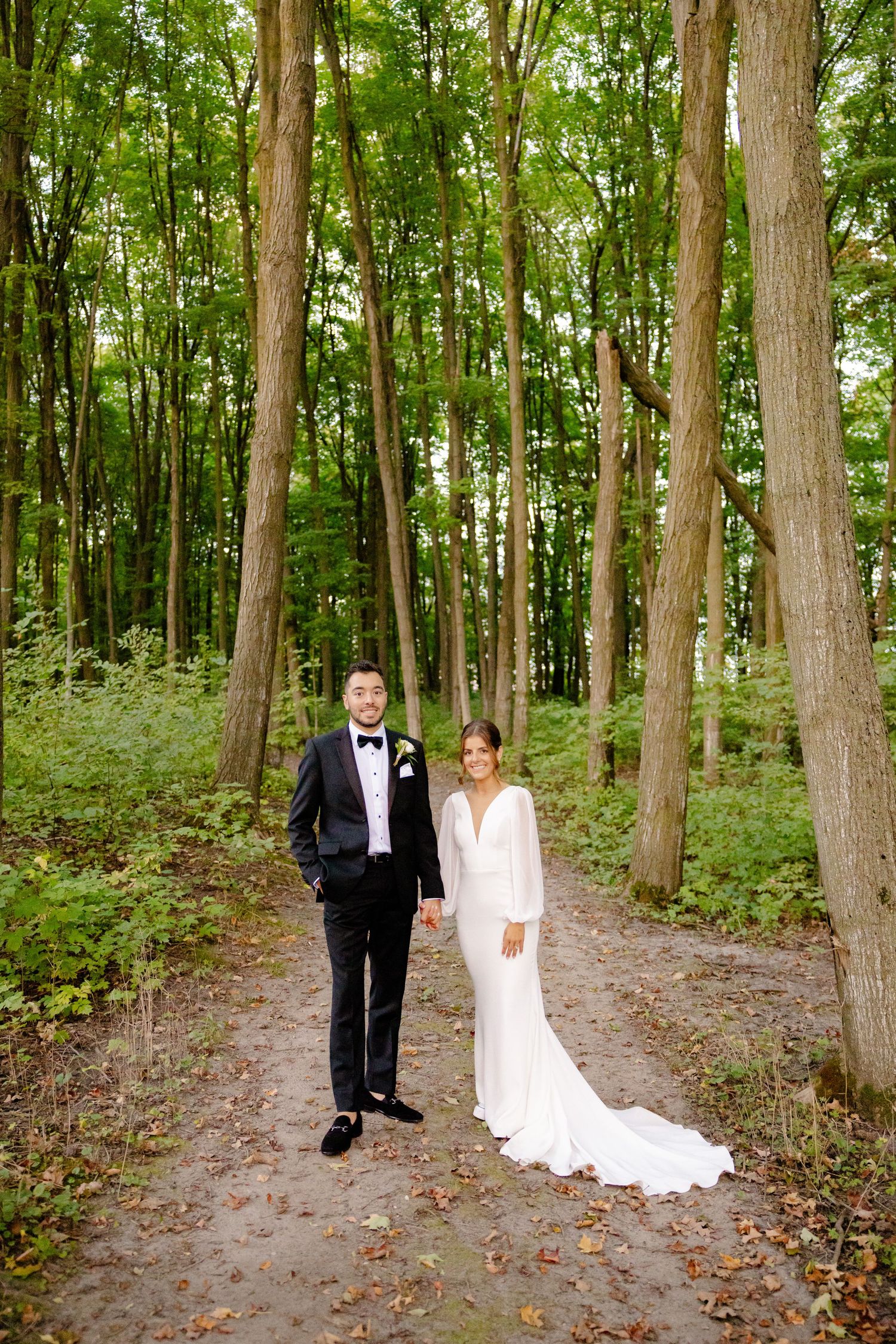 A bride and groom pose together on a woodland trail with sunlight filtering through the trees.