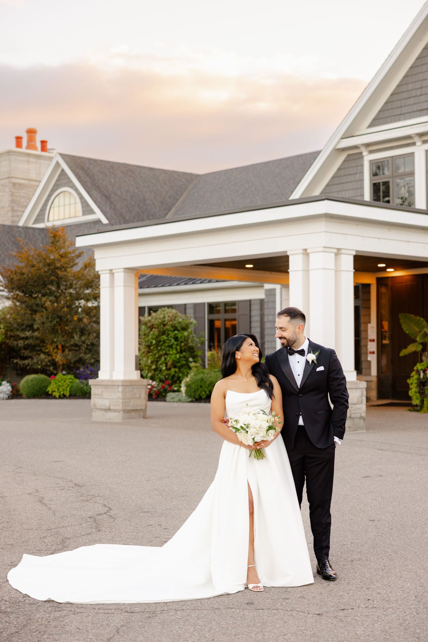 A wedding couple poses elegantly in front of a gray shingled venue with white columns at sunset.