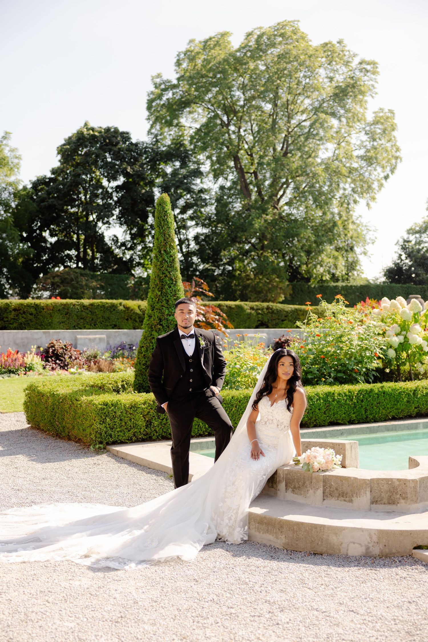 A couple poses on stone steps beside a garden pool with manicured hedges and towering trees.