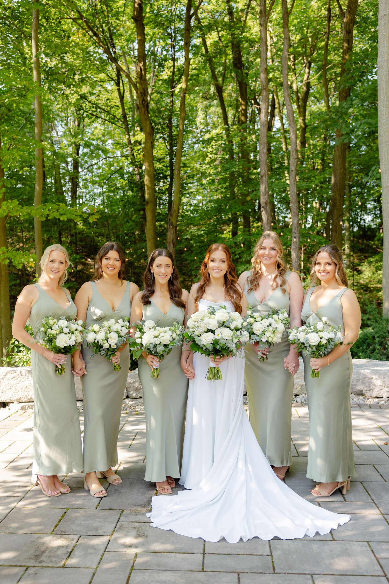 Wedding group in sage green dresses holding white bouquets on stone patio with forest backdrop.