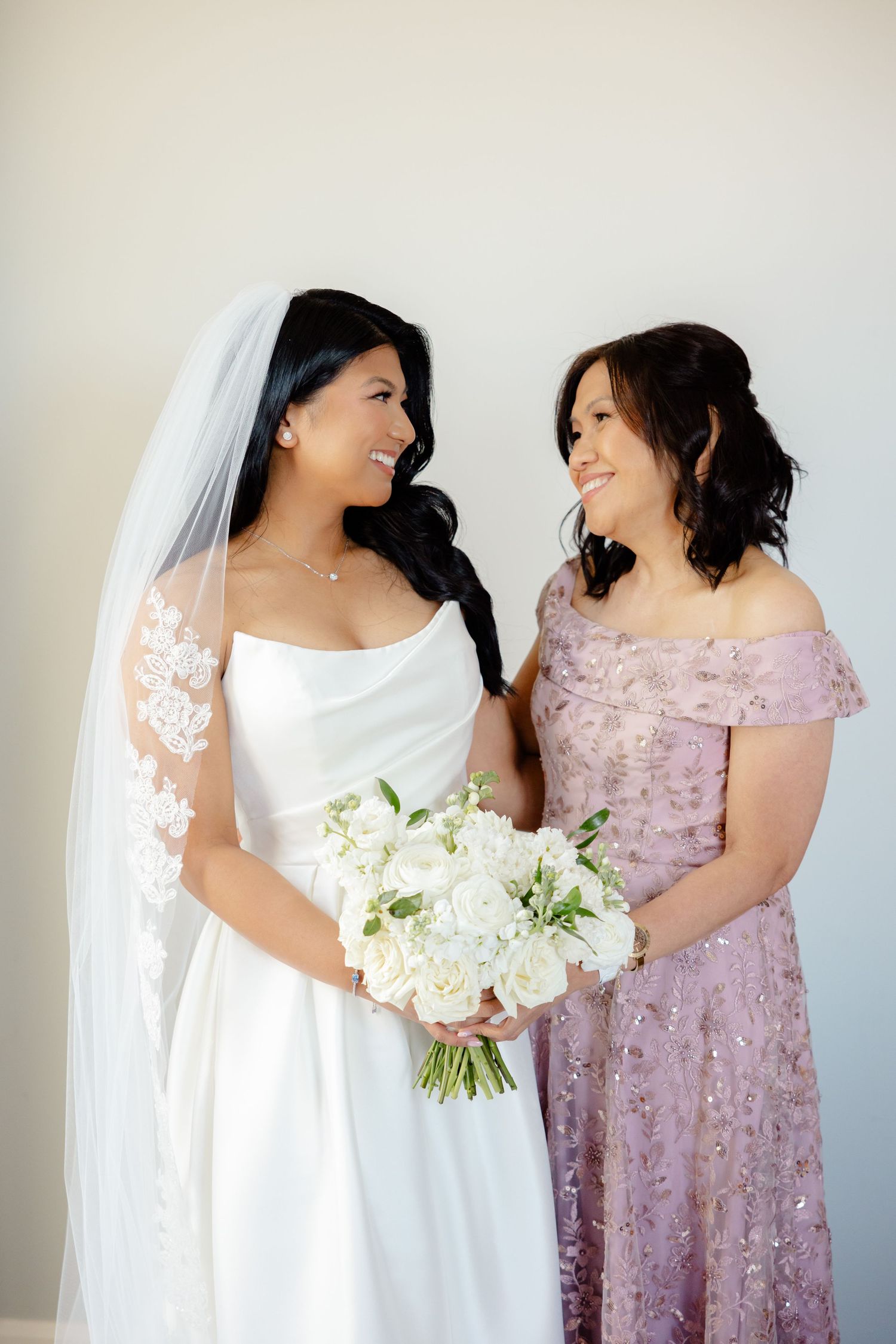 Tender moment shared between bride and woman in lilac dress while holding white bouquet.