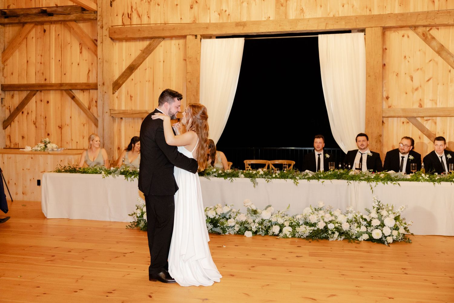 Couple shares their first dance in a rustic barn venue with elegant floral decorations and head table in background.