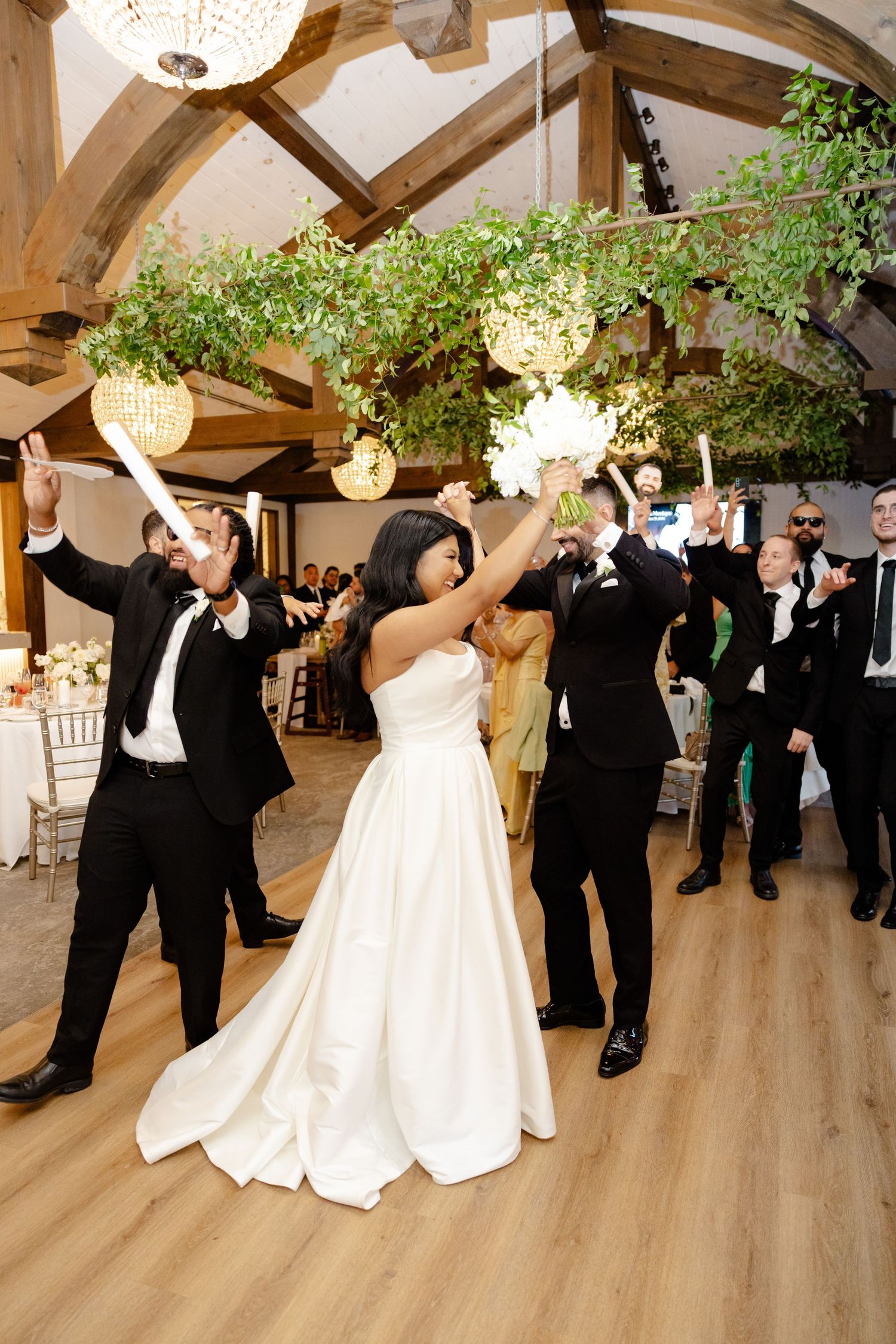 Wedding party celebrates with raised arms under crystal chandeliers and greenery decor.