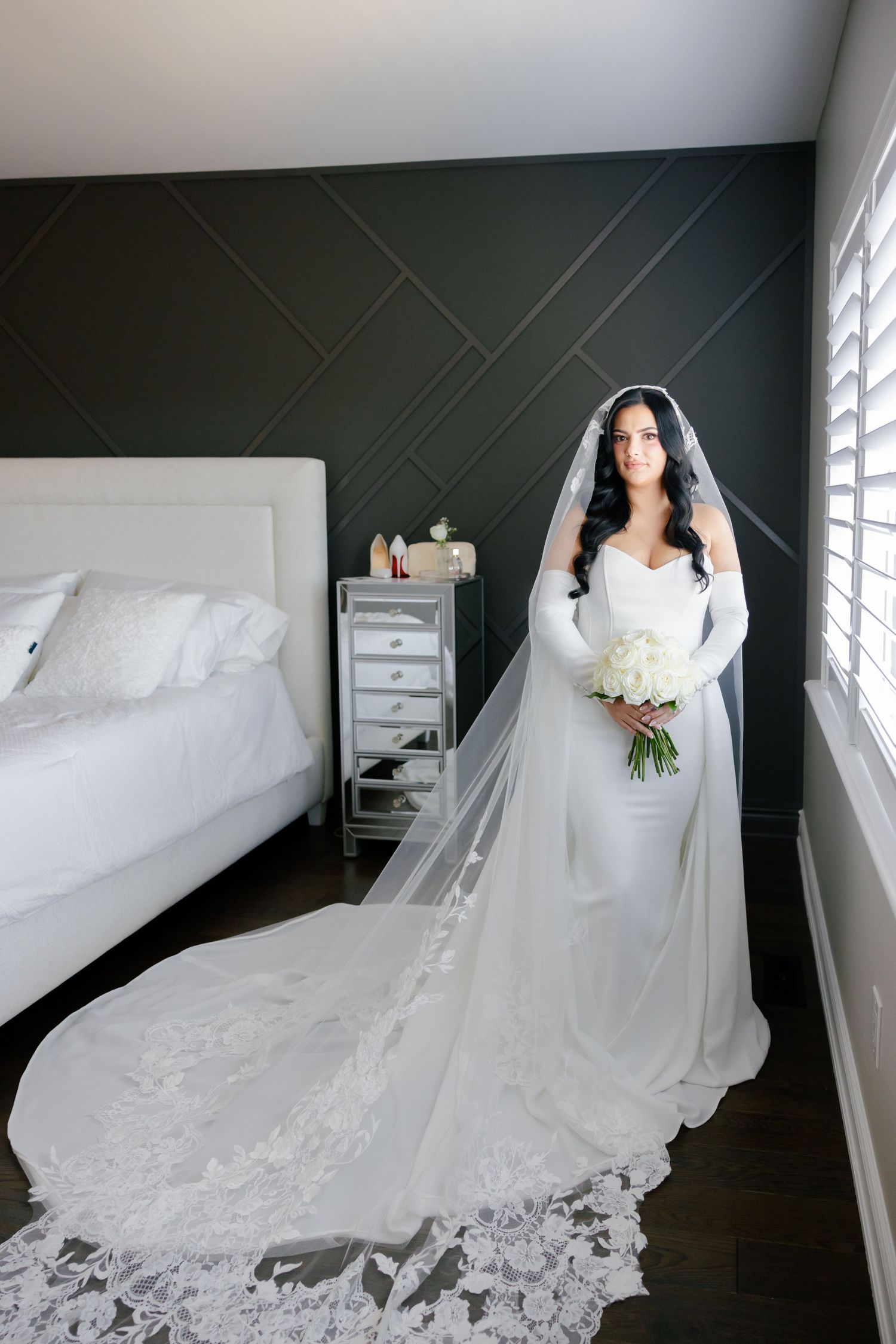 A stunning bridal portrait showcasing a long lace-trimmed veil and fitted white gown in a bedroom.