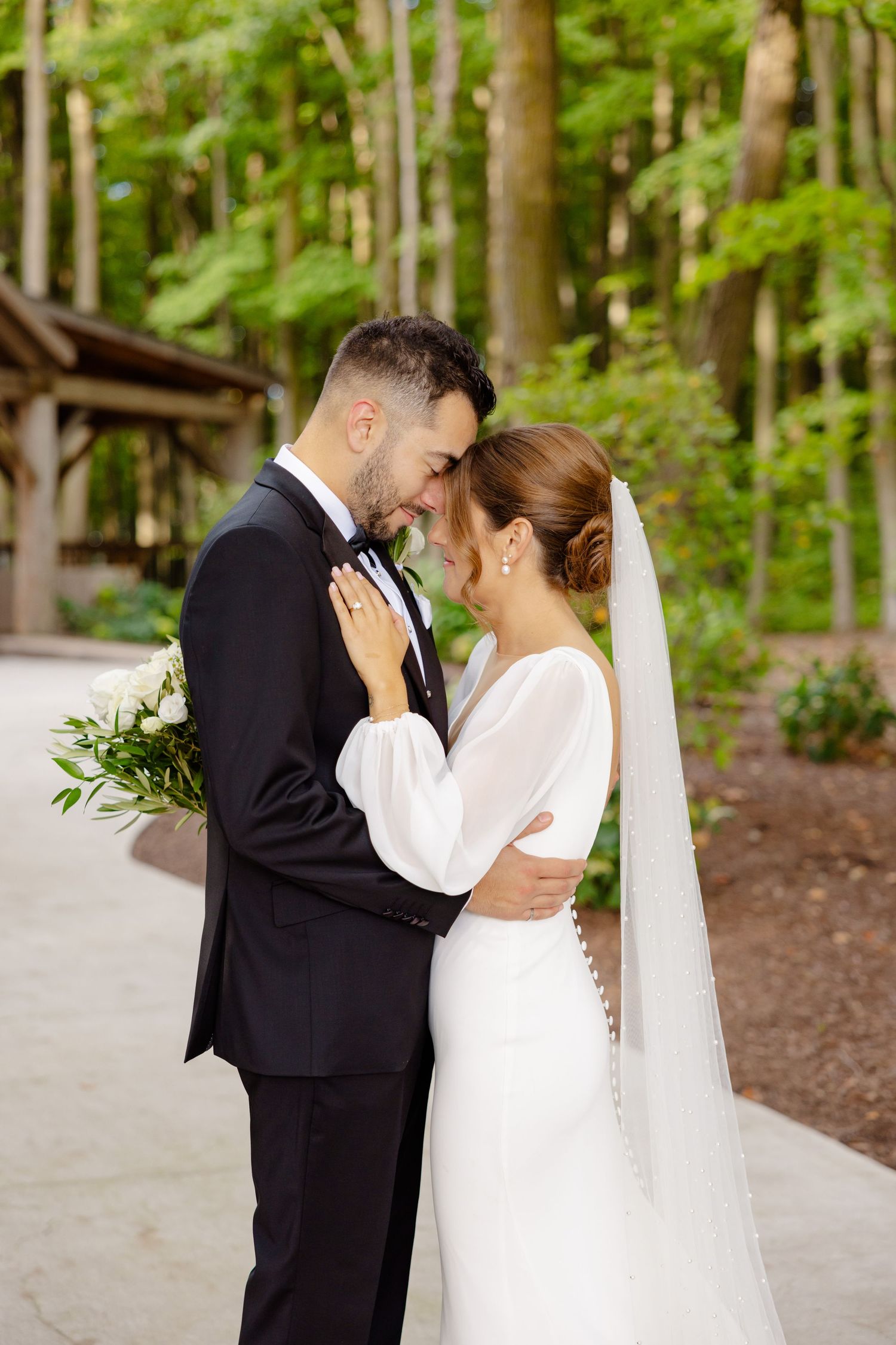 A romantic wedding moment between newlyweds embracing on a wooded path with a rustic pavilion in the background.