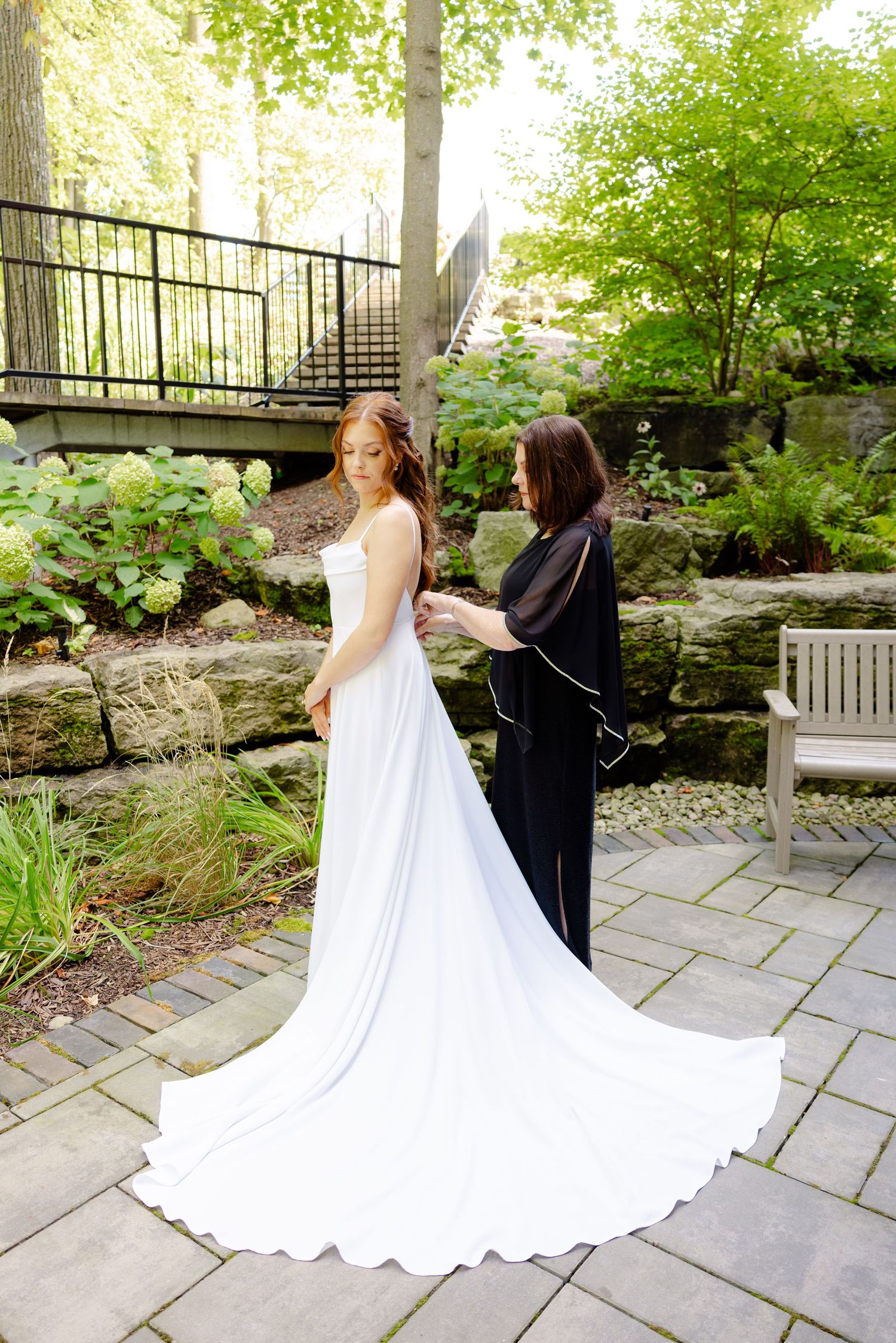 Wedding dress being adjusted by stylist in garden setting with stone pathway.