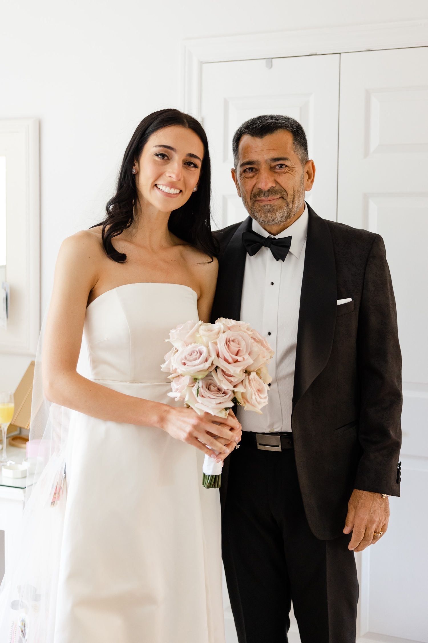 A bride in a strapless white gown holds a soft pink rose bouquet while posing with a companion in a tuxedo.