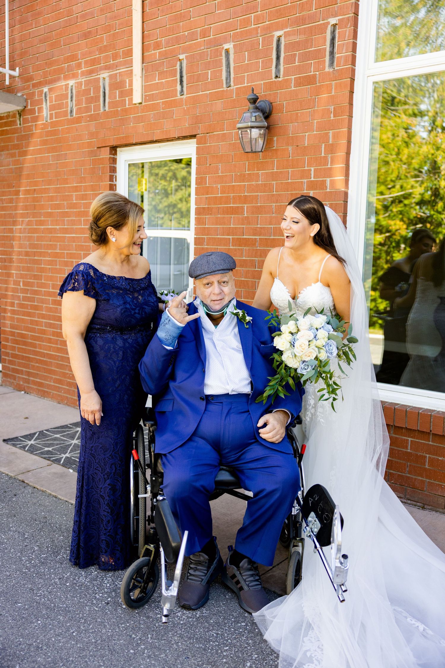 Wedding party poses by brick building with an elderly gentleman in a wheelchair wearing a blue suit.