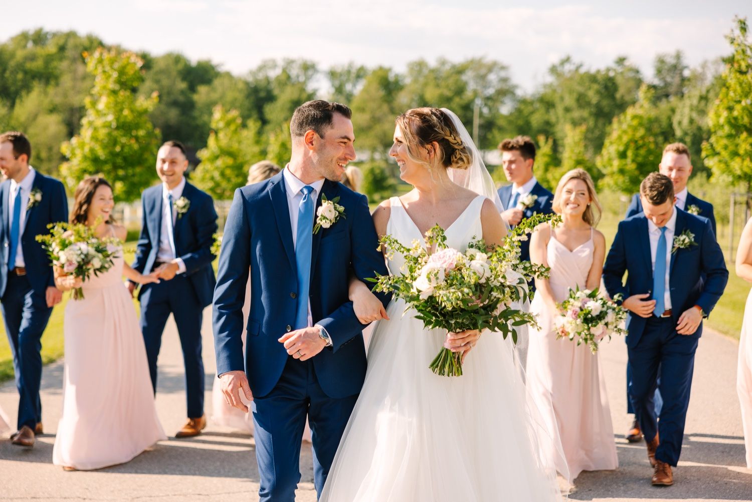 Wedding party walks together outdoors with bridesmaids in pink dresses and groomsmen in navy suits.