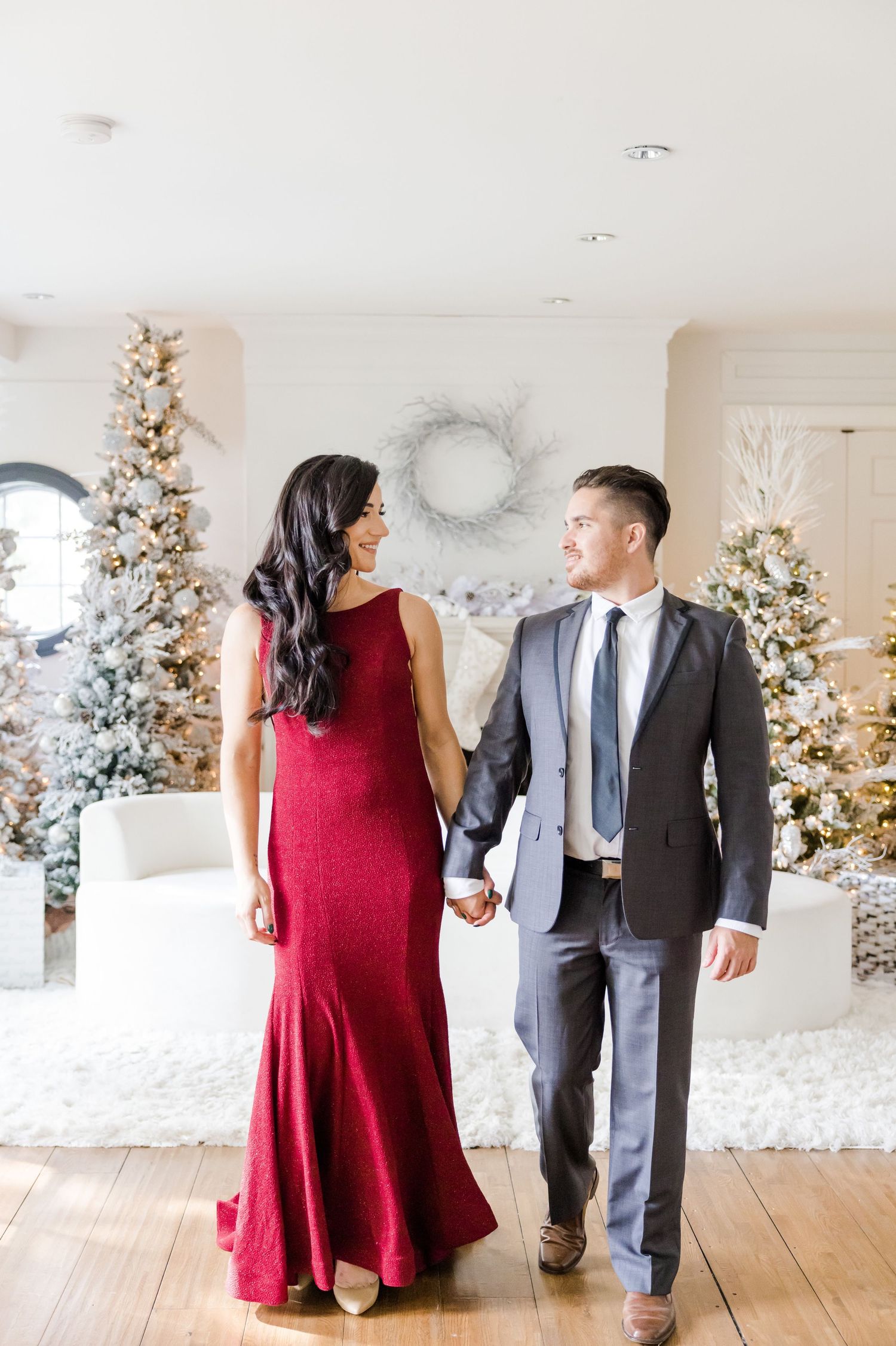 Couple walks hand in hand in formal attire against holiday decorated interior.
