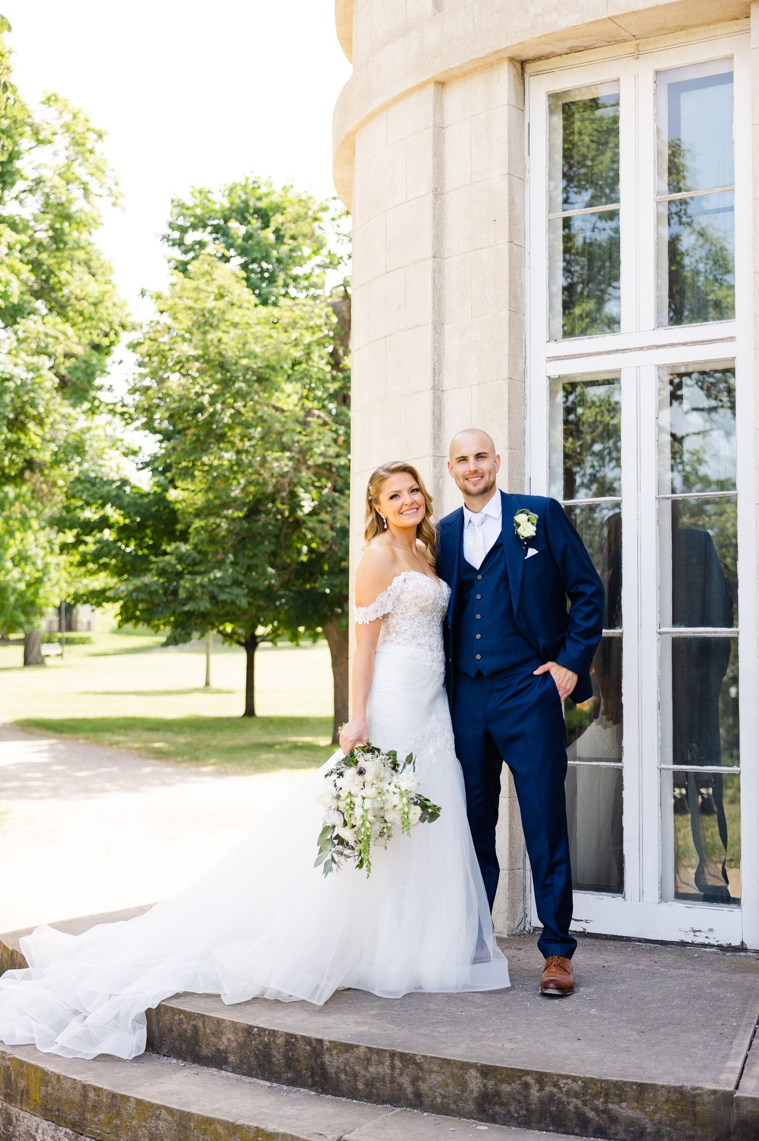 Newlyweds posing elegantly against classical white building with tall windows.