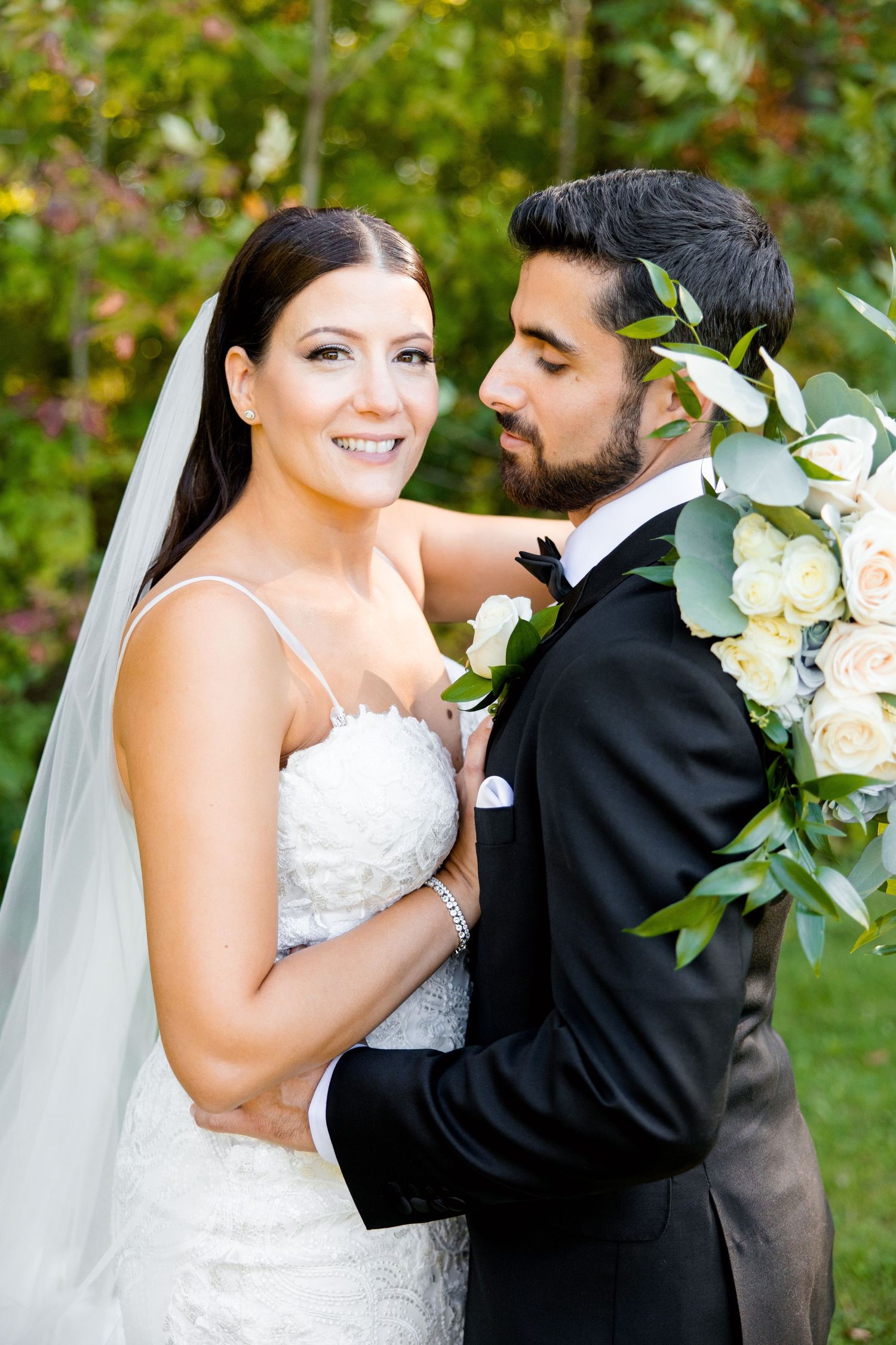 A romantic wedding portrait features a couple embracing with a stunning floral bouquet against a natural backdrop.