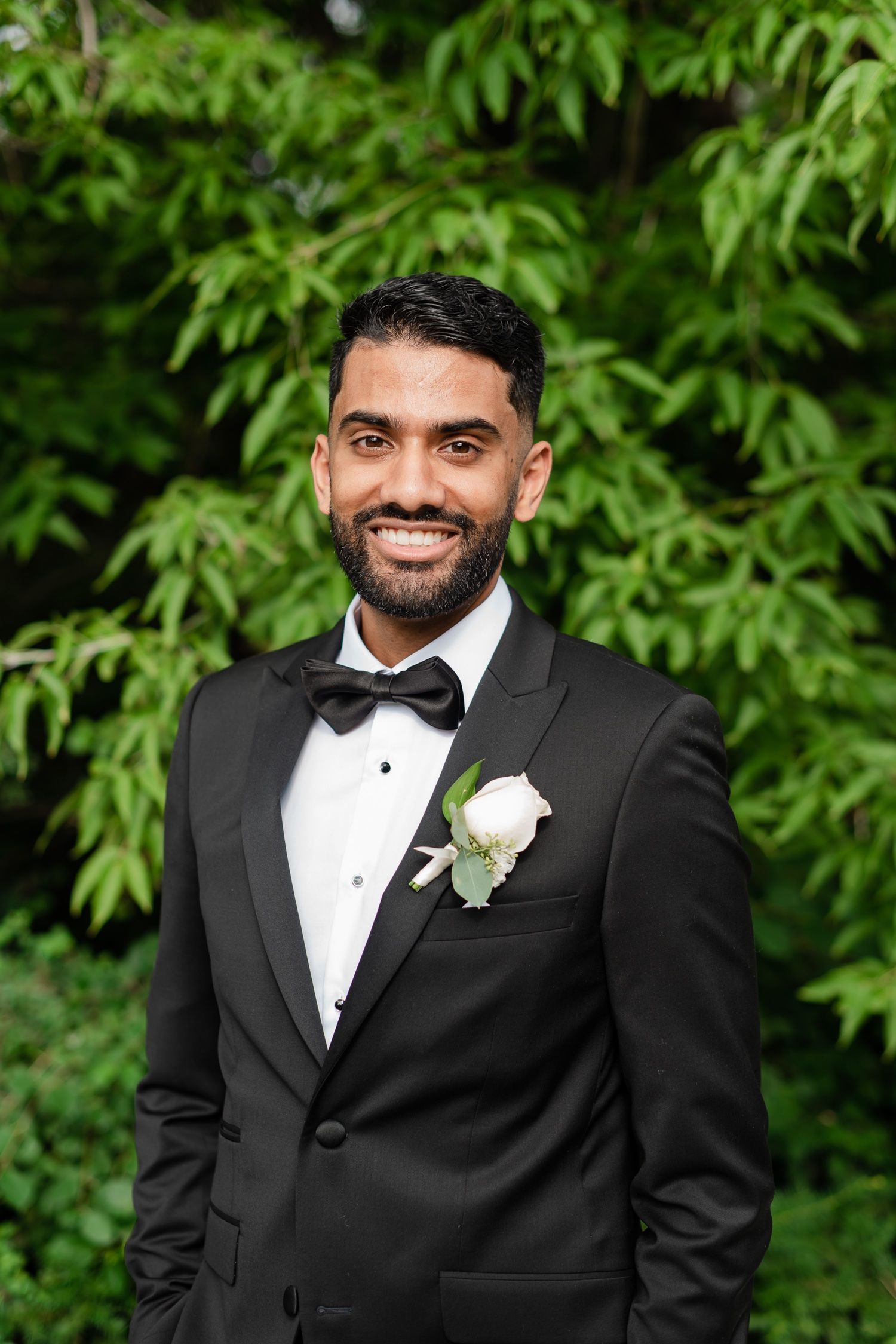 Wedding portrait with black tuxedo and white boutonniere against lush green background.