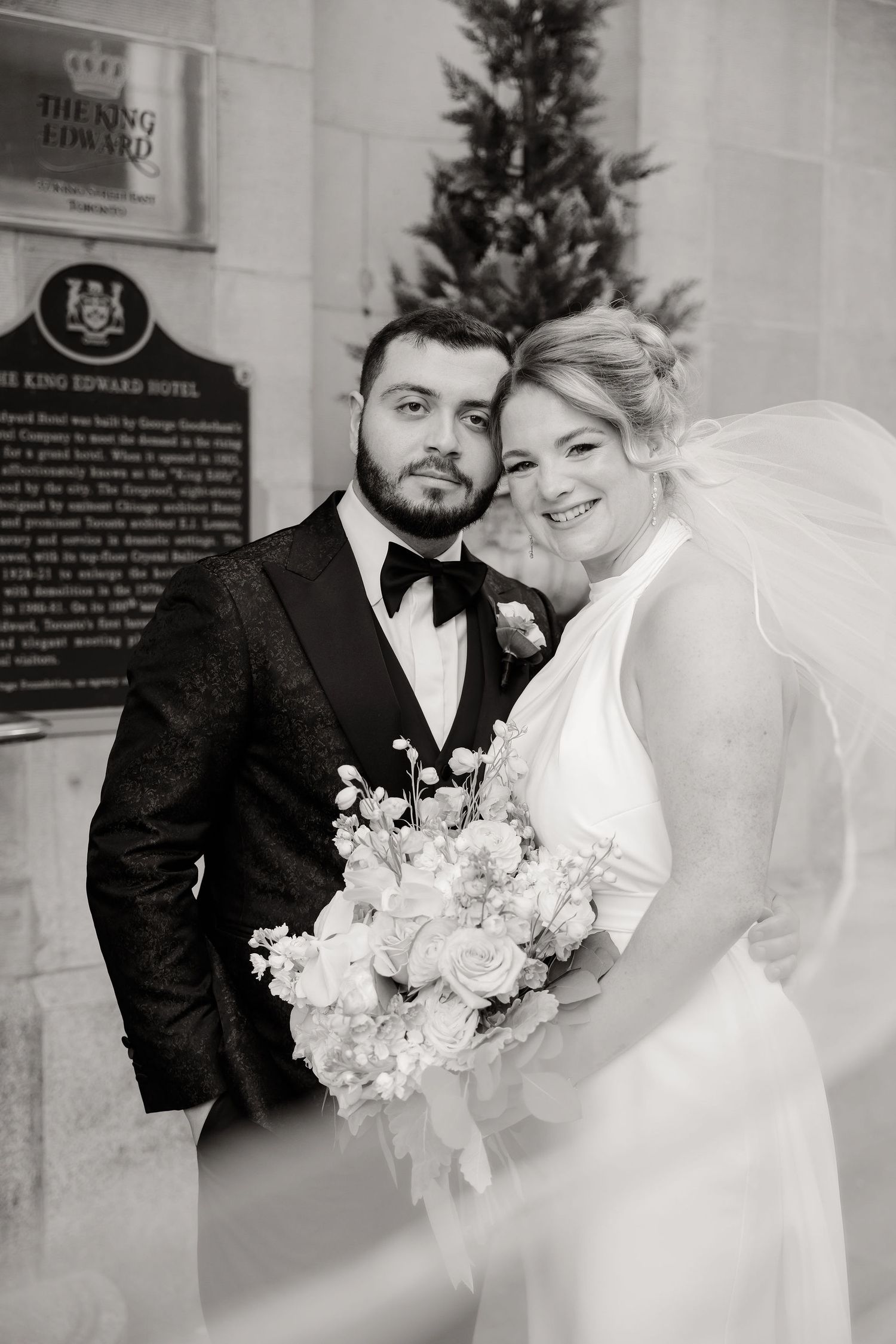 A black and white wedding portrait taken in front of a hotel sign with elegant floral bouquet.