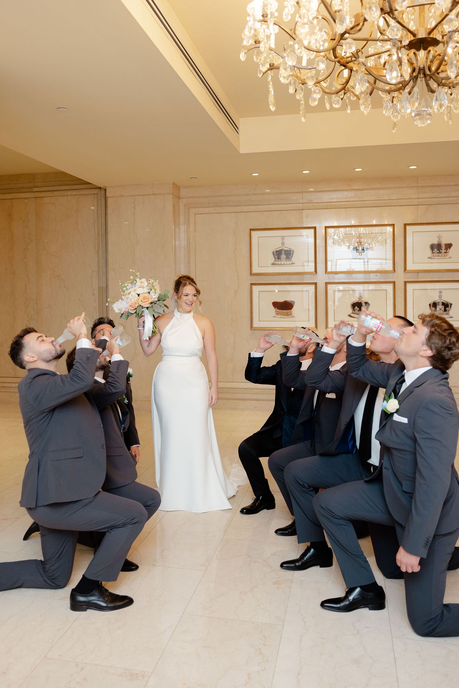 Wedding party performs synchronized kneeling pose in formal attire under crystal chandeliers.