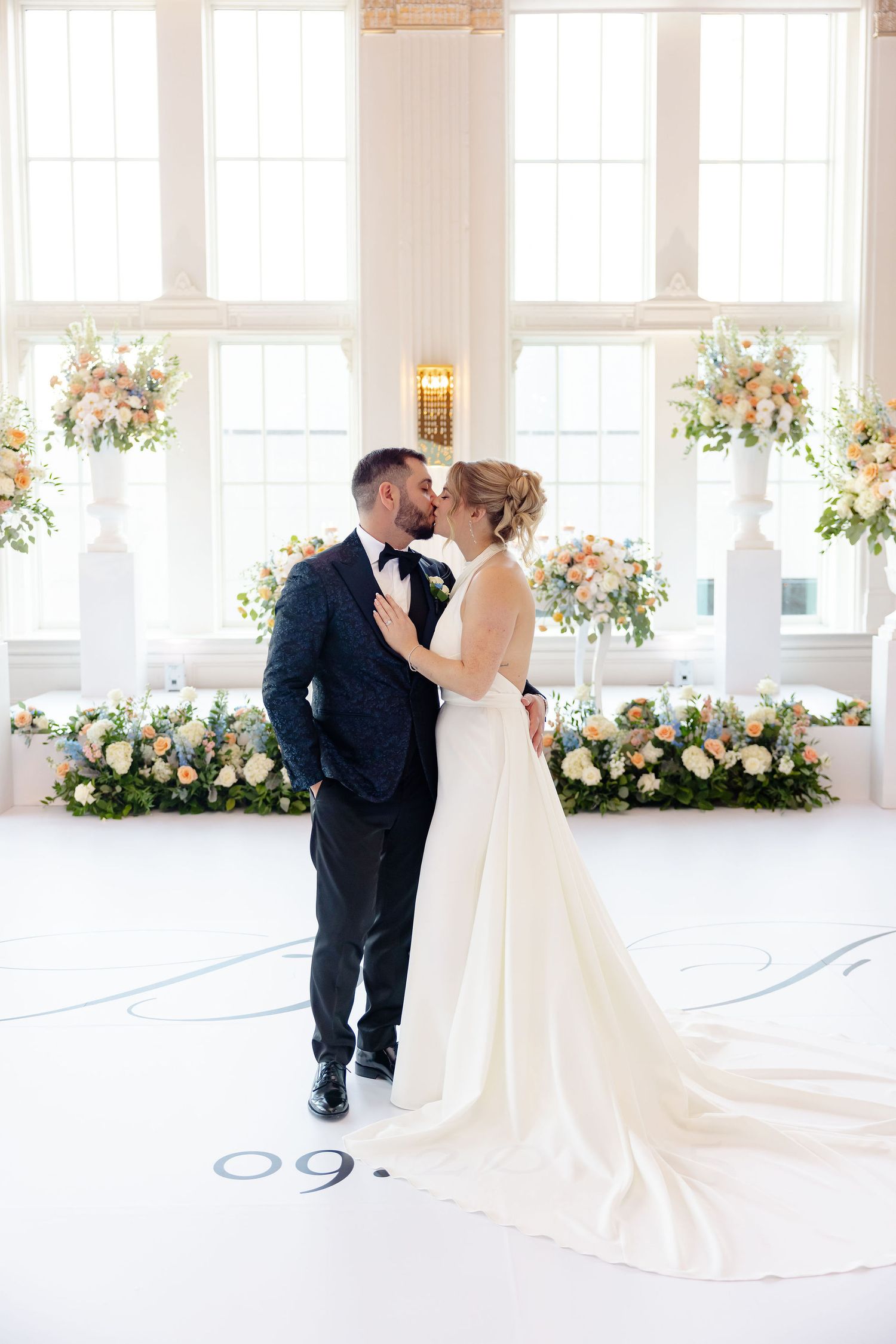 Romantic wedding moments captured in a bright white ballroom with floral decorations and large windows.