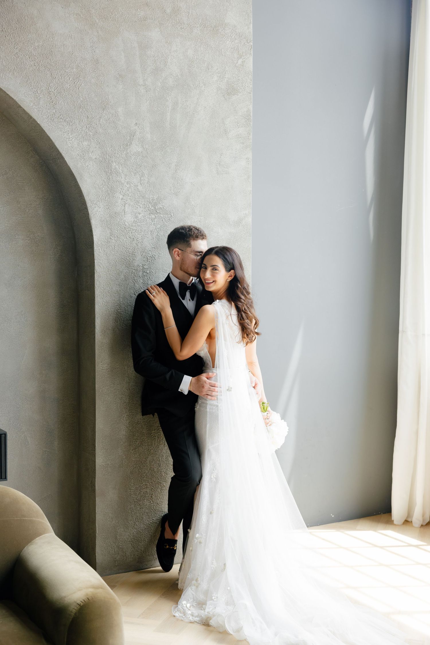 Newlywed couple in intimate moment against grey wall with natural window light.