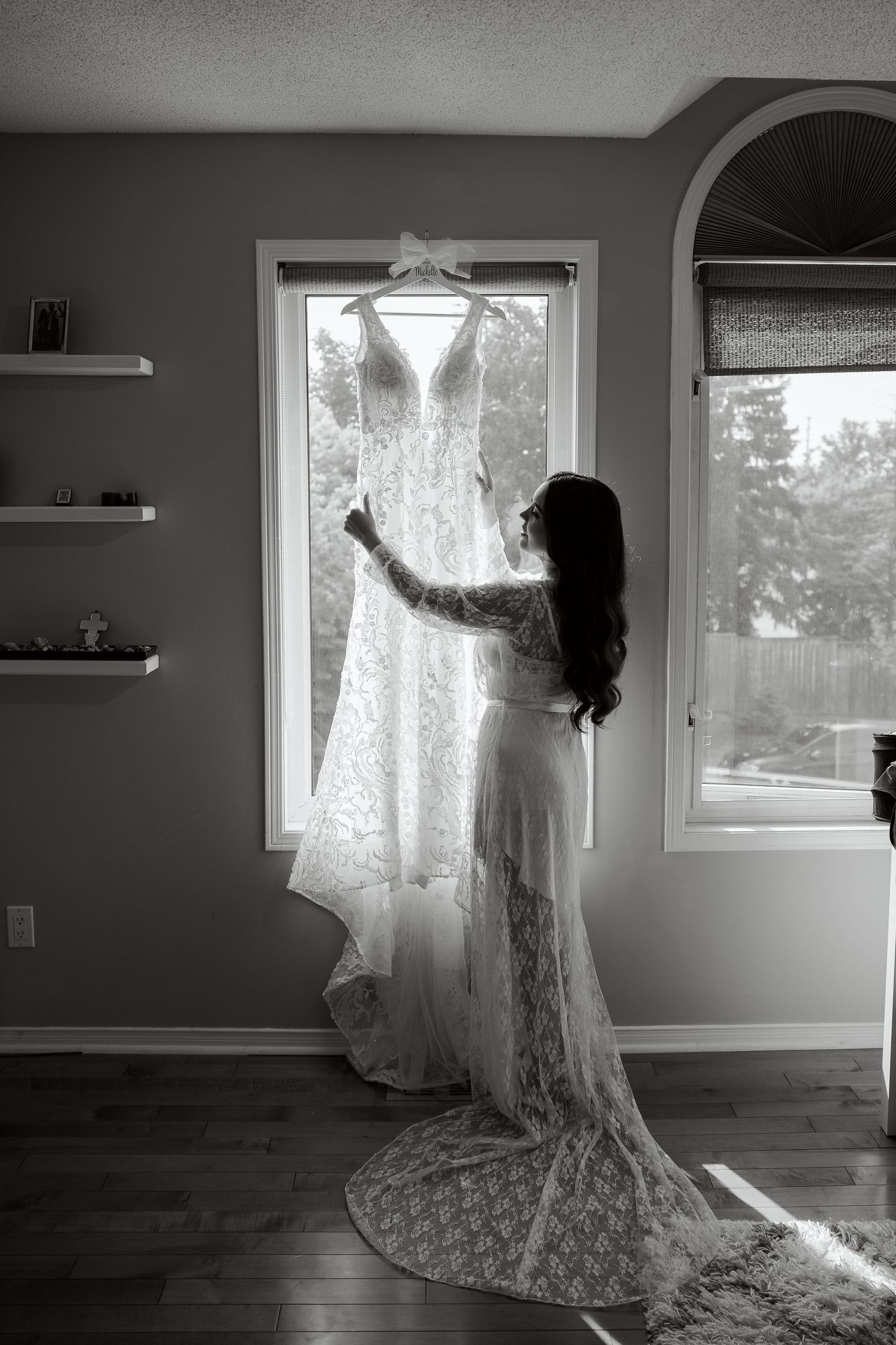 A silhouetted figure in a lace dress adjusts a white wedding gown hanging in front of a bright window.