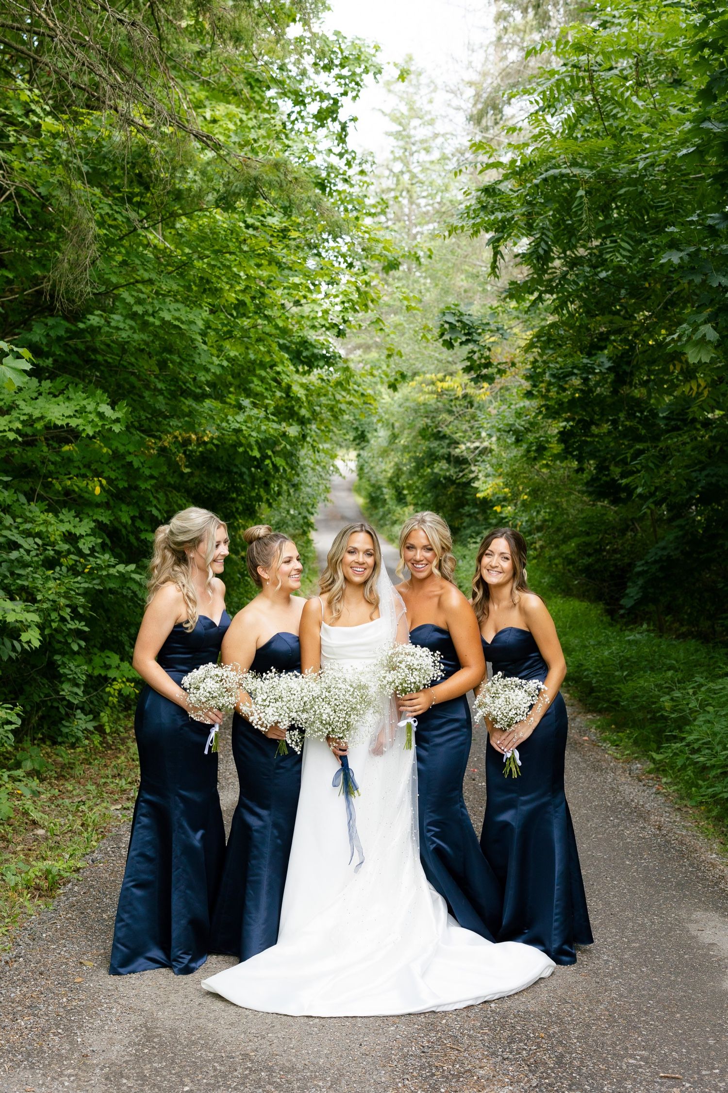 Wedding party featuring bridesmaids in navy gowns holding baby's breath bouquets on tree-lined path.