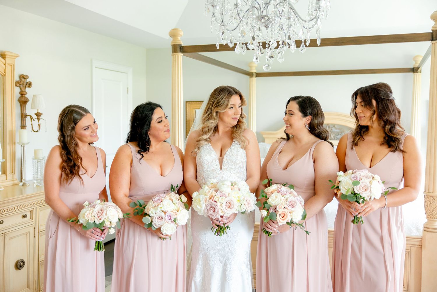 Joyful wedding party in matching pink dresses sharing candid moment in elegant bedroom setting.