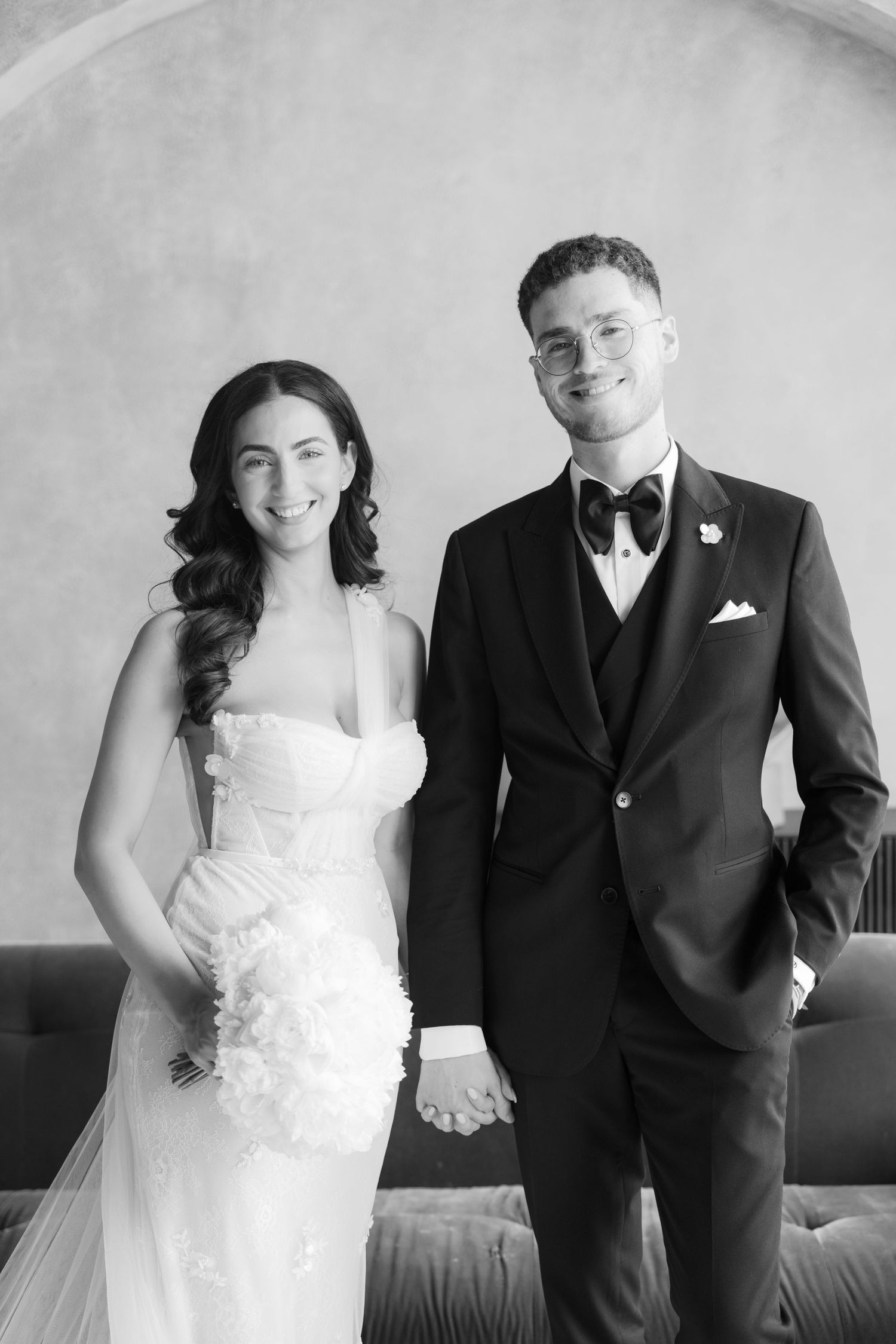Black and white portrait of newlyweds in formal attire standing in front of curved wall.