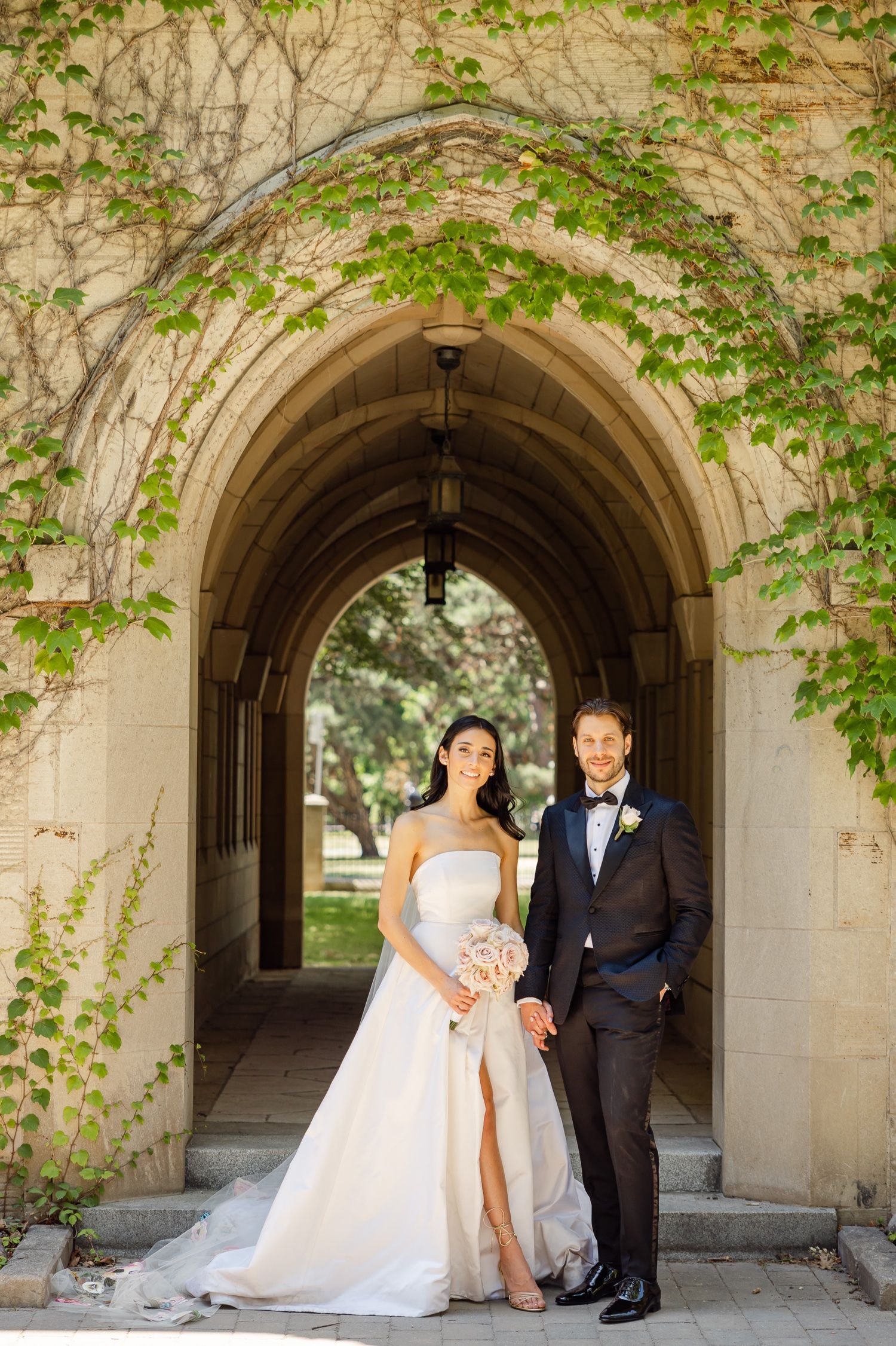 Couple poses in stone archway with climbing vines on university campus.